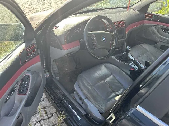 Wnętrze samochodu marki BMW widziane z otwartych drzwi kierowcy. Widoczna jest szara skórzana tapicerka foteli, kierownica z logo BMW, deska rozdzielcza oraz konsola środkowa z lewarkiem zmiany biegów. Elementy wykończenia wnętrza są podkreślone czerwonymi liniami.