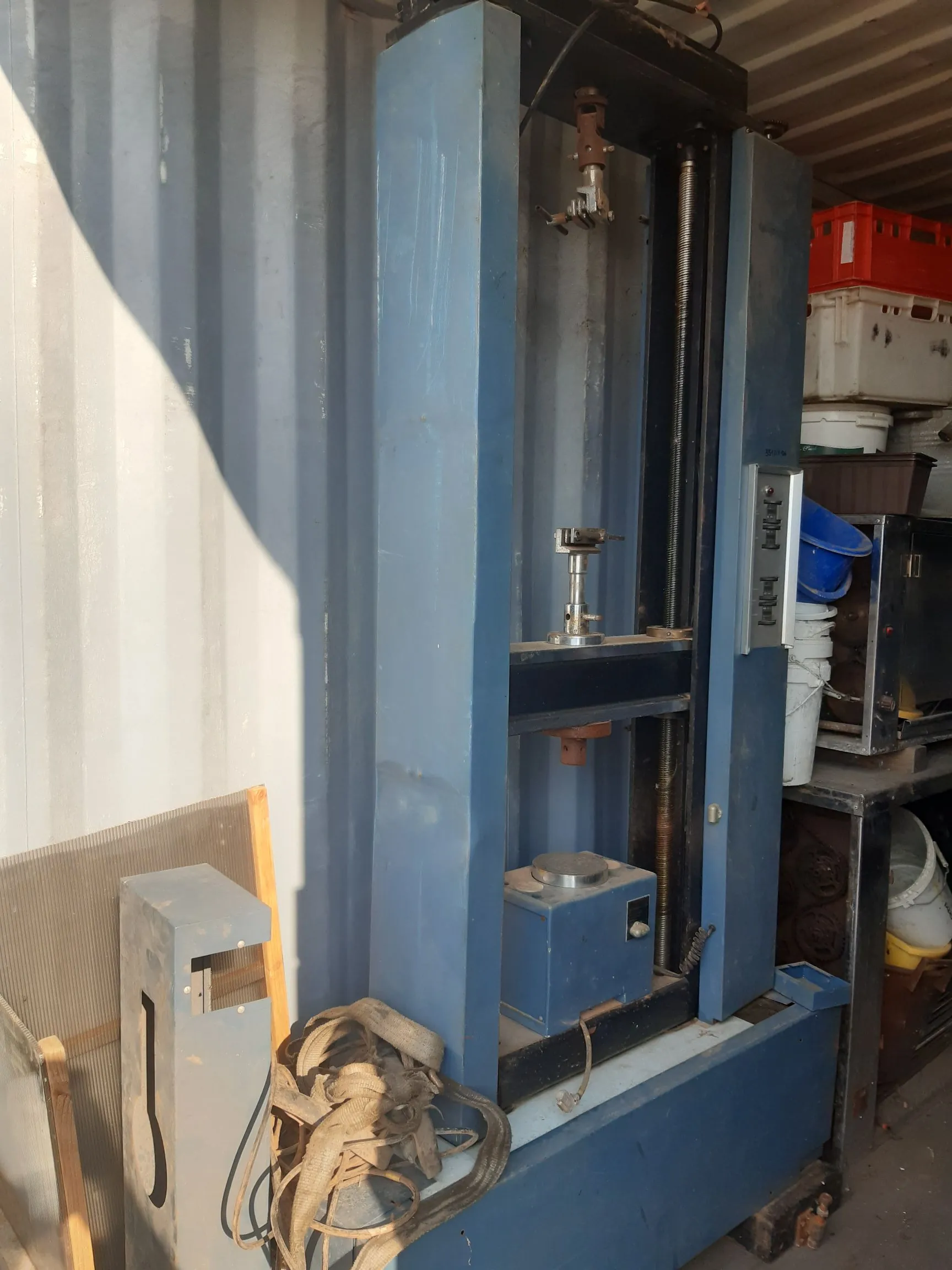 Niebieska, metalowa maszyna warsztatowa o pionowej konstrukcji, przypominająca prasę hydrauliczną lub urządzenie pomiarowe, ustawiona przy ścianie szarego kontenera. Maszyna posiada pionowe prowadnice, siłownik oraz podstawę. Obok urządzenia leży zwinięty, jasnobrązowy pas transportowy. W tle widoczne są regały z plastikowymi pojemnikami i sprzętem warsztatowym.
