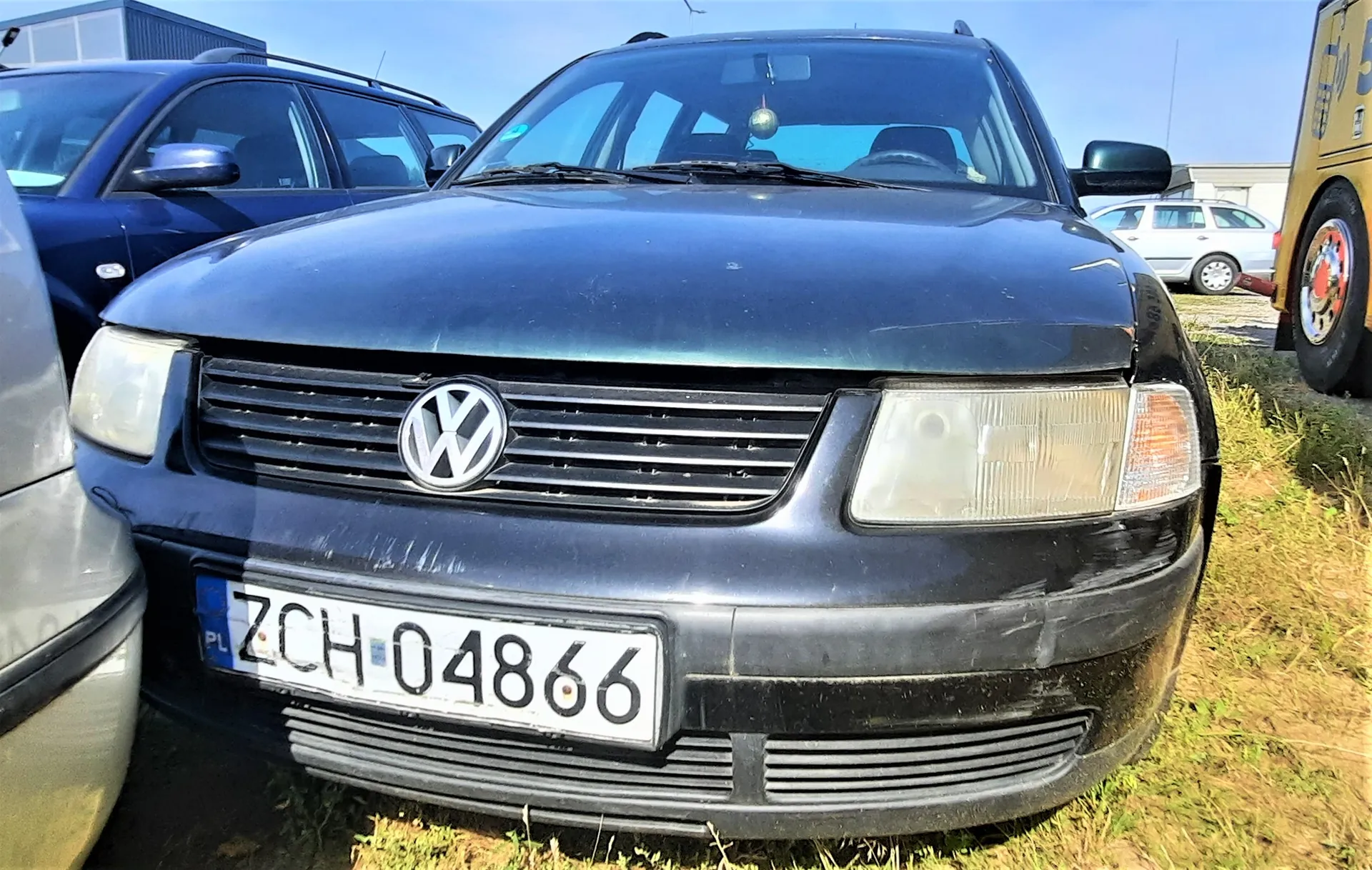 Widok z przodu ciemnoniebieskiego samochodu marki Volkswagen. Pojazd posiada polską tablicę rejestracyjną o numerze ZCH 04866. Samochód stoi na trawiastym terenie, obok niego widoczne są inne pojazdy: niebieski samochód po lewej stronie oraz biały i żółty pojazd w tle po prawej stronie.