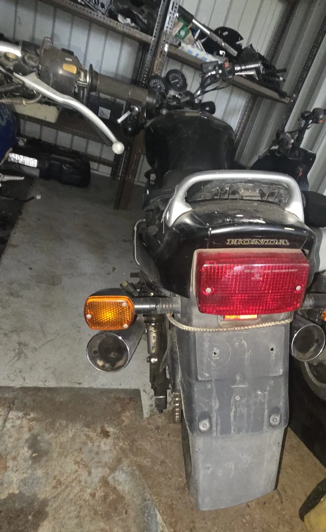 Tylne widok czarnego motocykla marki Honda w garażu. Widoczne czerwone światło tylne, pomarańczowe kierunkowskazy, metalowy uchwyt na bagaż oraz napis 'HONDA' na pokrywie. W tle metalowe półki z narzędziami, częściami mechanicznymi i betonowa podłoga z plamami.