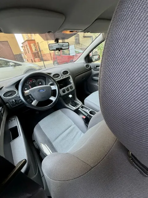 Wnętrze samochodu marki Ford widziane z perspektywy tylnego siedzenia. W kadrze znajduje się szary materiałowy fotel pasażera w pierwszym planie, kierownica z logo Ford, deska rozdzielcza oraz przedni fotel. Przez przednią szybę widoczny jest fragment białego samochodu, czerwony pojazd oraz budynki mieszkalne.