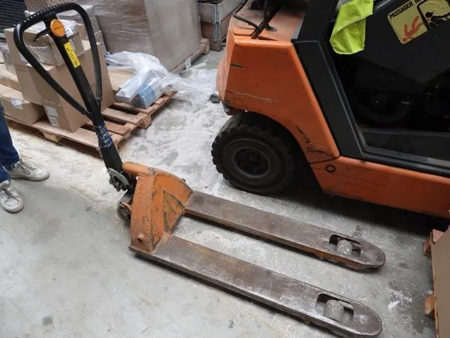 Pomarańczowy wózek paletowy stojący na betonowej posadzce w magazynie. Wózek ma dwa metalowe widelce skierowane w prawą stronę oraz czarny uchwyt do sterowania. W tle widoczny jest fragment pomarańczowego wózka widłowego oraz paleta z kartonami i folią ochronną.