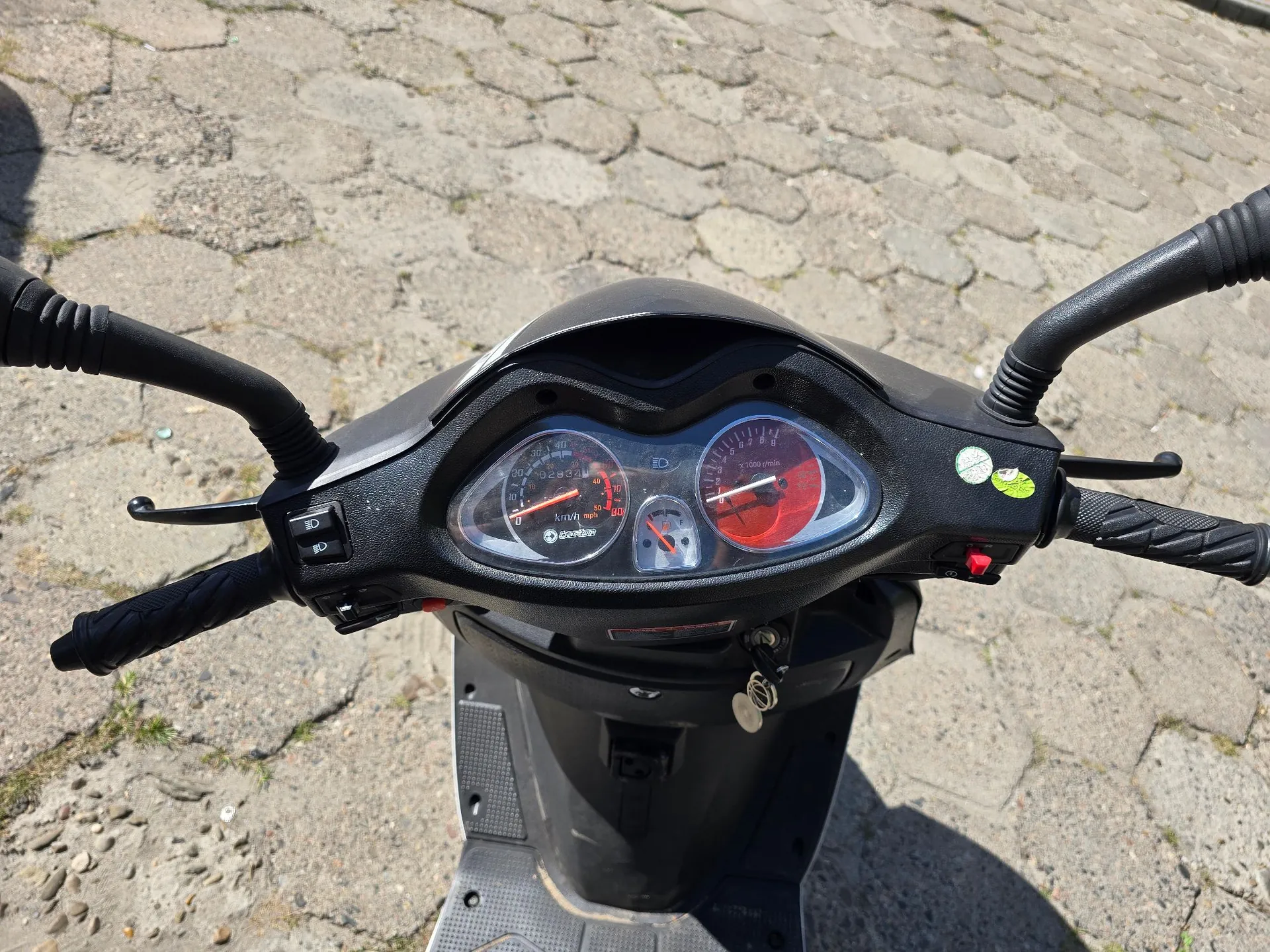 Widok z perspektywy kierowcy na kierownicę i deskę rozdzielczą czarnego skutera. Na desce rozdzielczej znajdują się dwa okrągłe wskaźniki: lewy z prędkościomierzem i obrotomierzem oraz prawy z czerwonym tłem. Po lewej stronie kierownicy widoczny jest przełącznik świateł z ikoną żarówki. Skuter stoi na szarej, kostce brukowej.