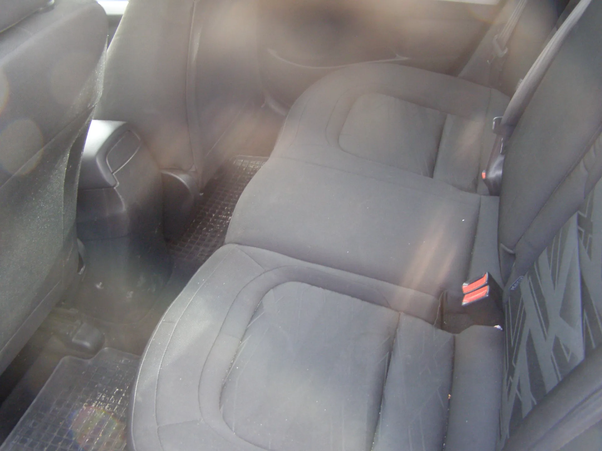 Wnętrze samochodu z widokiem na tylne siedzenia. Szare siedzenia tapicerowane tkaniną, widoczne pasy bezpieczeństwa z czerwonymi elementami zapięć. Na podłodze czarne gumowe dywaniki. Fragmenty siedzeń i drzwi są lekko zabrudzone. Obraz jest nieco rozmyty, ale wszystkie elementy są wizualnie rozpoznawalne.