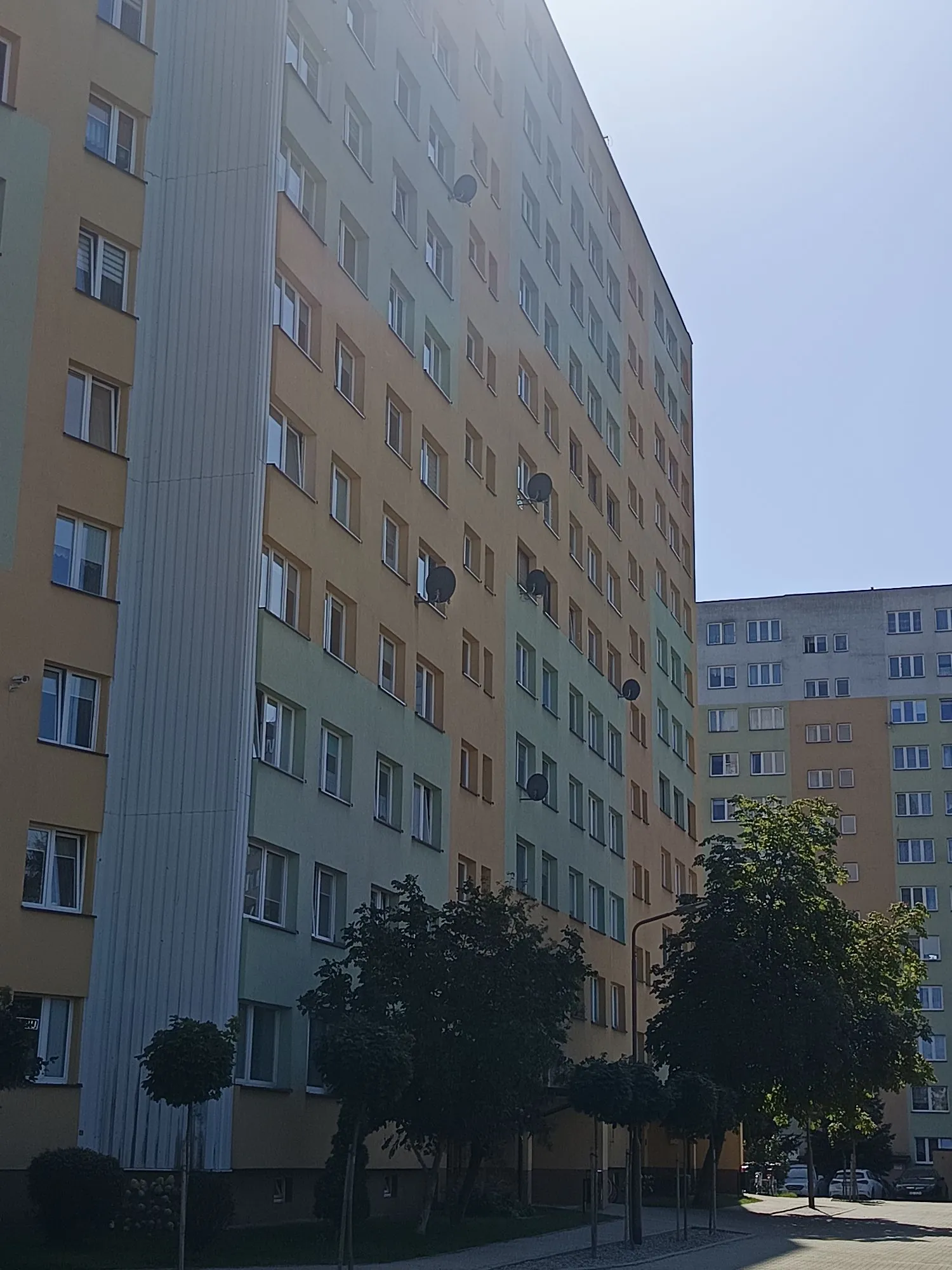 Wysoki blok mieszkalny z wielkiej płyty o elewacji w kolorach żółtym, zielonym i szarym. Budynek posiada liczne okna oraz zamontowane na ścianach anteny satelitarne. Przed blokiem rosną zielone drzewa i krzewy. W tle widoczny jest fragment drugiego, podobnego budynku mieszkalnego pod jasnym, bezchmurnym niebem.