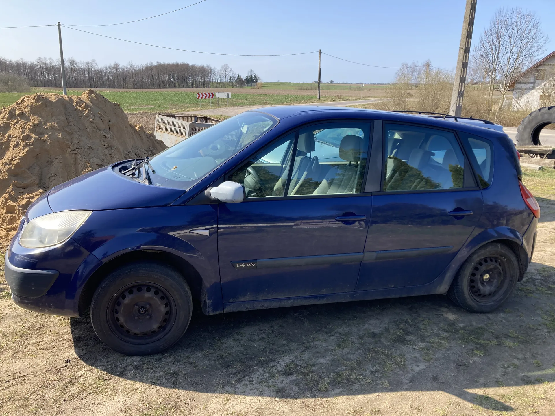 Niebieski samochód osobowy typu hatchback z napisem "1.4 16V" na boku, zaparkowany na brudnej, ziemistej powierzchni. W tle widoczna duża kopca ziemi, pola, drzewa, słupy energetyczne oraz znak drogowy z czerwono-białymi strzałkami. Po prawej stronie obrazu znajduje się duży opona i budynek. Niebo jest jasne, bezchmurne.