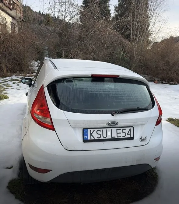 Biały samochód marki Ford z tyłu, z tablicą rejestracyjną KSU LE54 (PL) umieszczoną na środku. Samochód stoi na śnieżnej powierzchni z widocznymi plamami trawy. W tle suche krzewy, drzewa bez liści, fragment budynku po lewej stronie oraz wzgórze z lasem. Na tylnej szybie widoczny wycieraczka, a na bagażniku logo marki. Niebo częściowo zachmurzone.