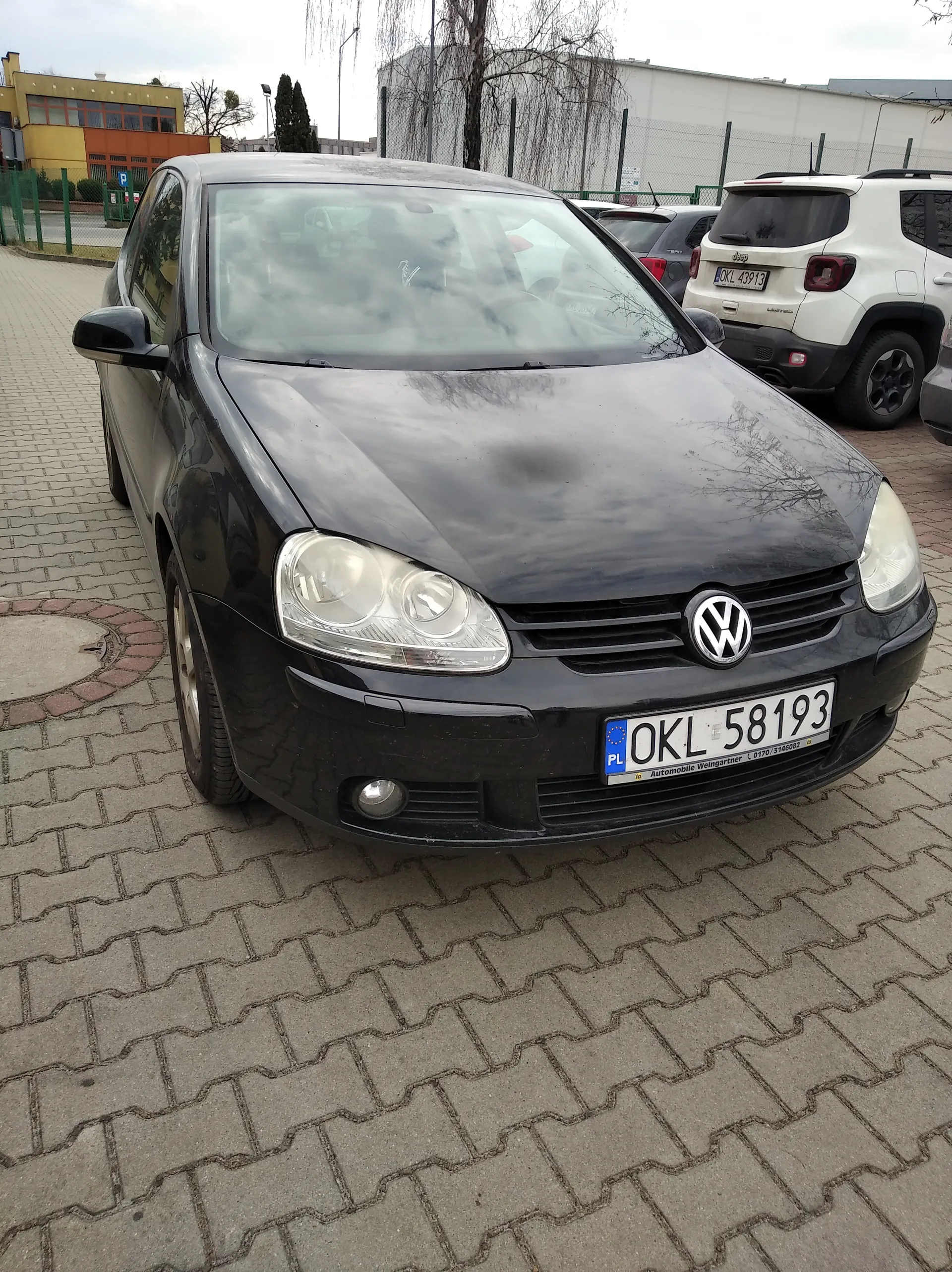 Czarny samochód Volkswagen z tablicą rejestracyjną PL OKL 58193 zaparkowany na brukowanym placu parkingowym. W tle biały SUV z tablicą OKL 43913, budynki, drzewa i ogrodzenie. Niebo zachmurzone.