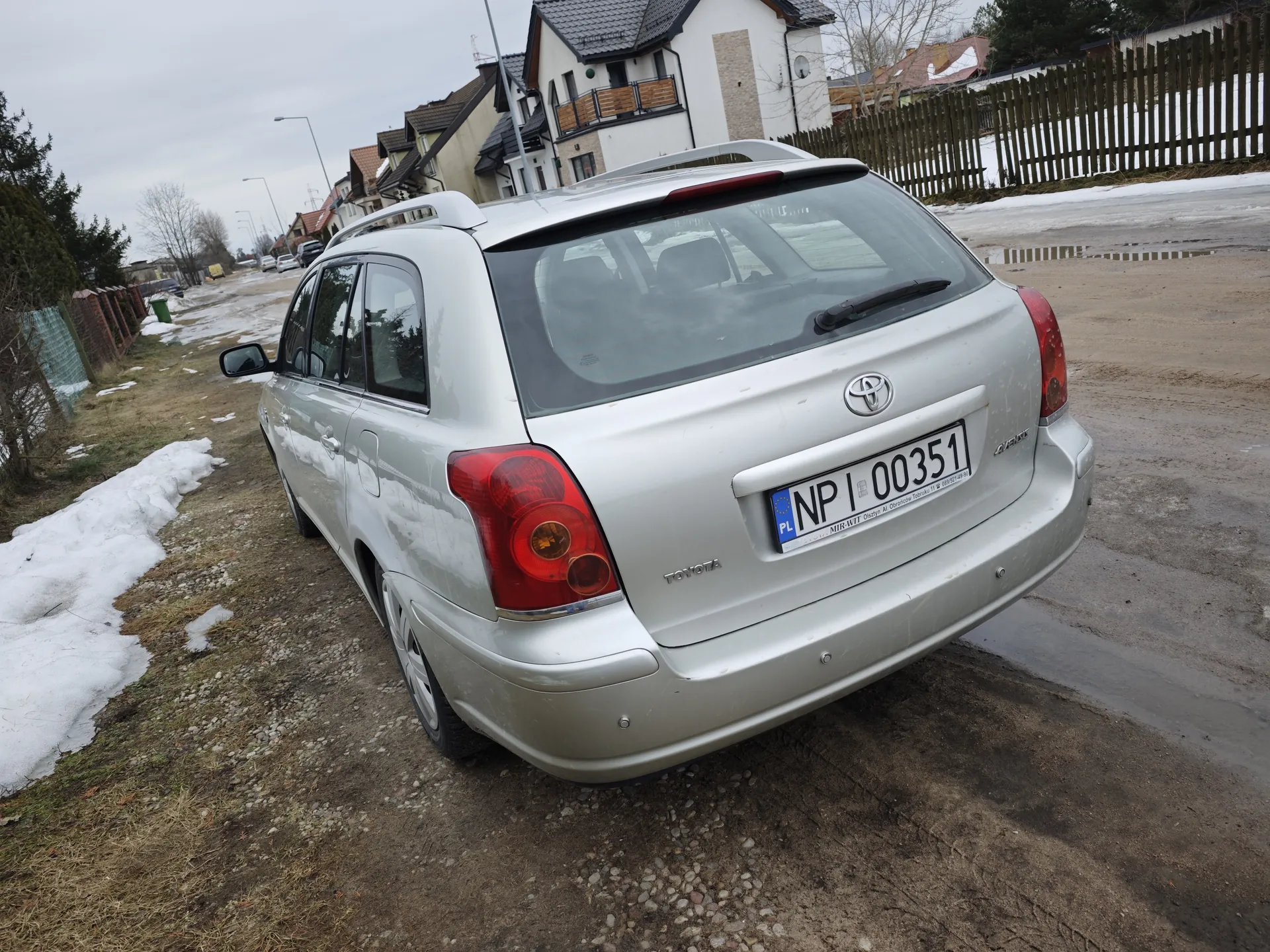 Srebrny samochód osobowy marki Toyota, widok od tyłu, z tablicą rejestracyjną PL NP 00351. Pojazd zaparkowany na drodze z żwirem i ziemią, po lewej stronie widoczne resztki śniegu. W tle domy mieszkalne z dachami, drewniany płot oraz drzewa bez liści. Niebo jest zachmurzone.