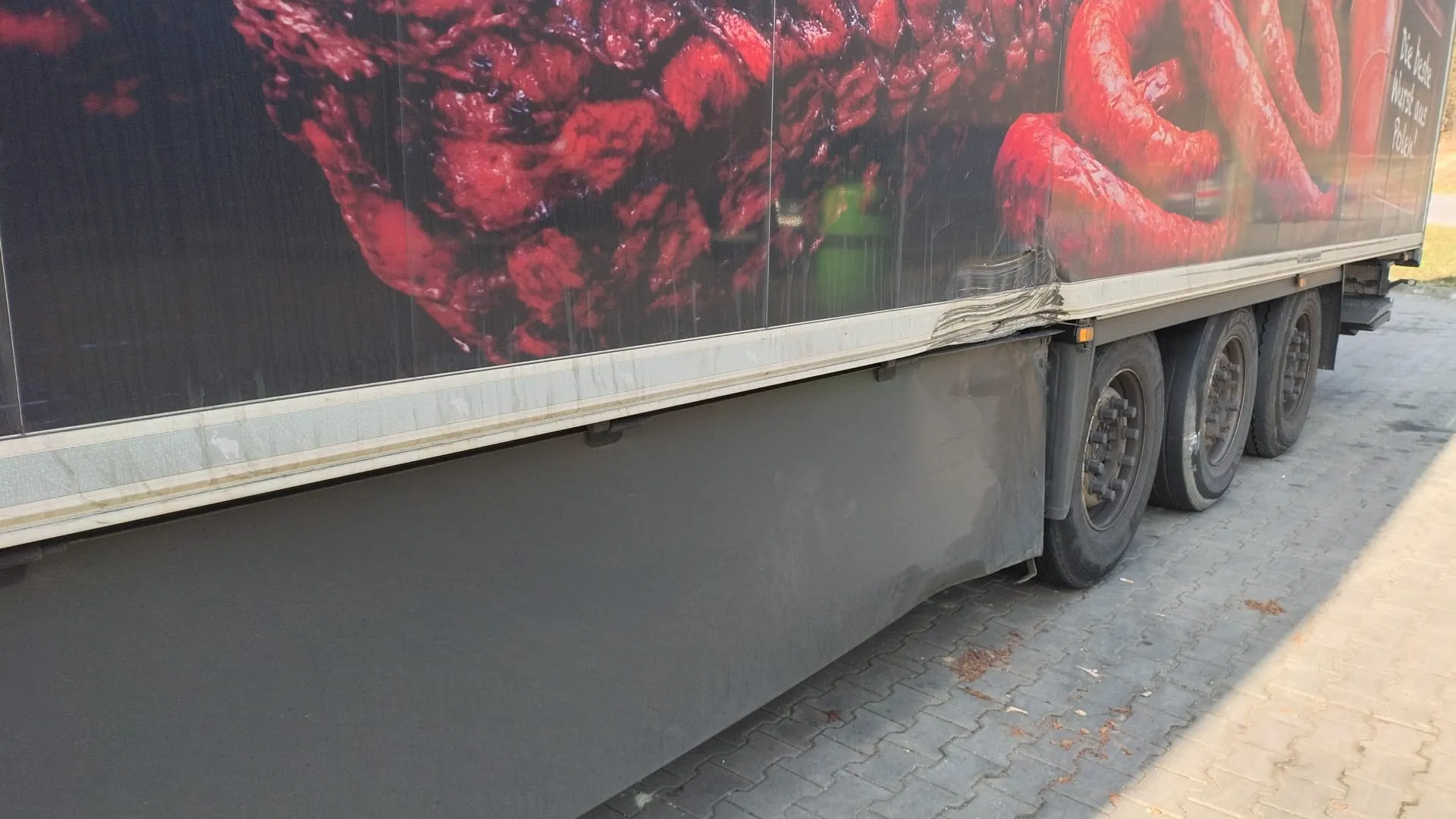 Boczna część przyczepy ciężarowej z dużą reklamą przedstawiającą czerwone mięso i kiełbasy. Widoczny napis w języku niemieckim: 'Die beste Wurst aus Polen!'. Przyczepa stoi na brukowanej powierzchni, widoczne są koła oraz uszkodzona krawędź nadwozia. W tle zielony obiekt.