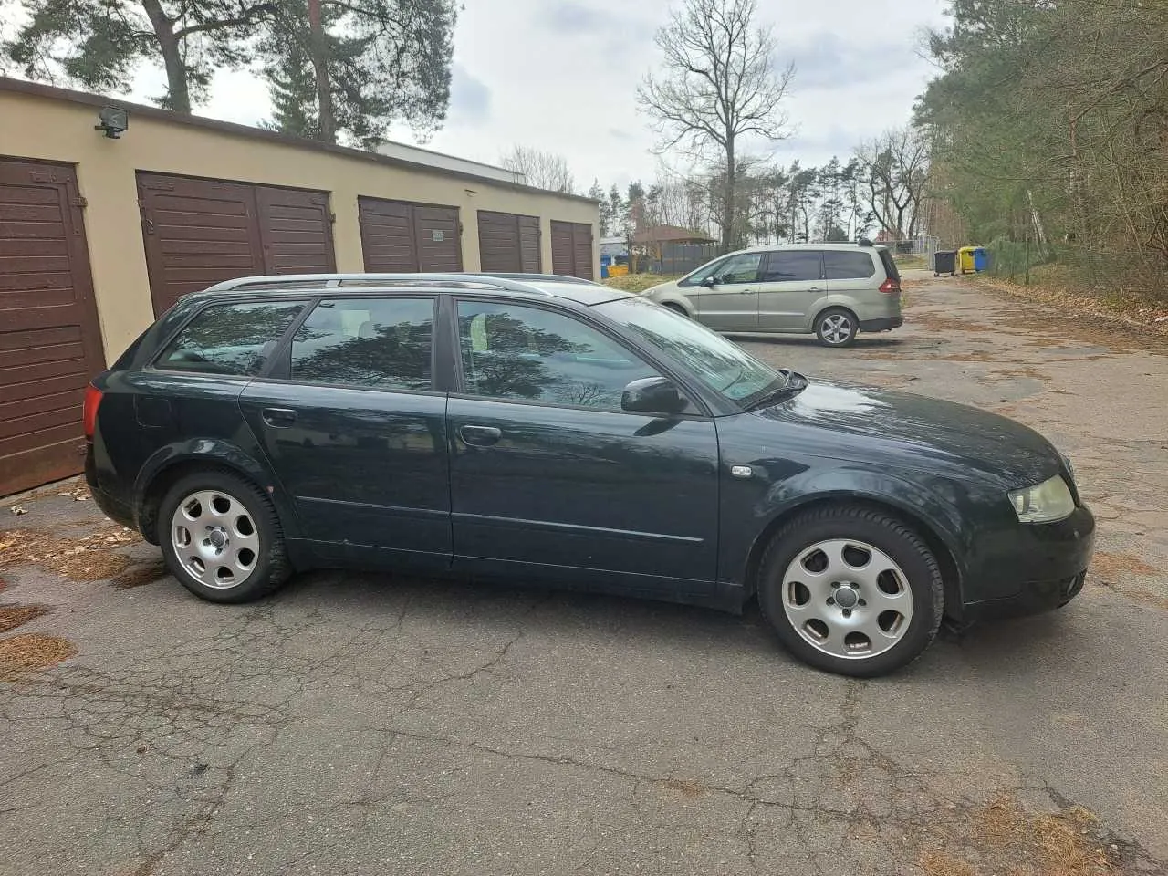 Ciemnozielony samochód typu kombi zaparkowany na pękniętym asfalcie. W tle budynek z brązowymi garażami, srebrny minivan, drzewa i pojemniki na śmieci. Niebo jest zachmurzone.