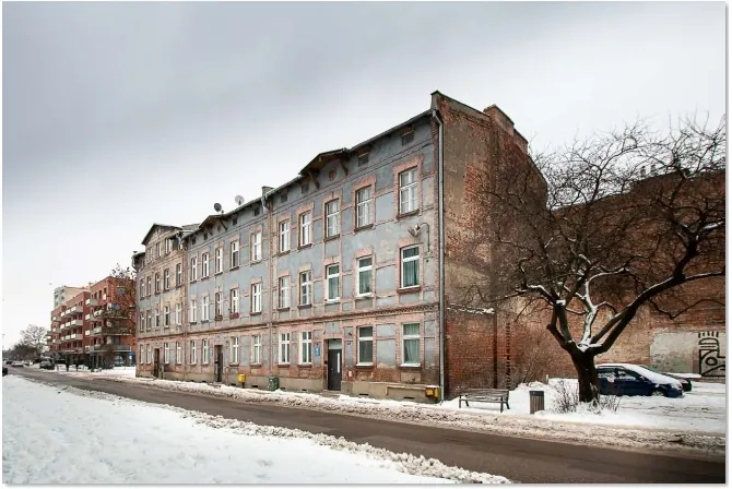 Zdjęcie przedstawiające wielopiętrową, starą kamienicę z cegły z niebiesko-szarym tynkiem, który w wielu miejscach odpada, odsłaniając czerwoną cegłę. Budynek posiada liczne, regularnie rozmieszczone okna. Po prawej stronie znajduje się bezlistne drzewo oraz zaparkowany ciemny samochód. Przed budynkiem przebiega asfaltowa droga, a pobocza i teren wokół są pokryte śniegiem. W tle po lewej stronie widoczny jest nowszy, wielorodzinny budynek mieszkalny. Niebo jest zachmurzone i szare.