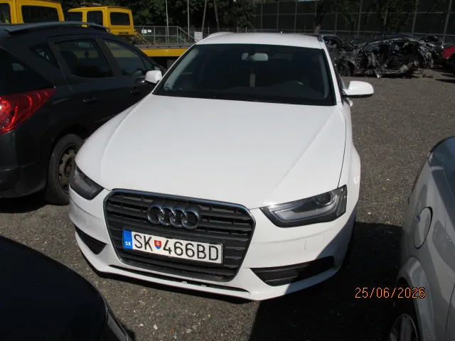 Biały samochód marki Audi zaparkowany na żwirowym placu. Widoczny jest przód pojazdu z rejestracją SK 466BD. Po lewej stronie znajduje się czarny samochód, a po prawej fragment srebrnego auta. W tle widoczne są żółte pojazdy oraz wrak samochodu.
