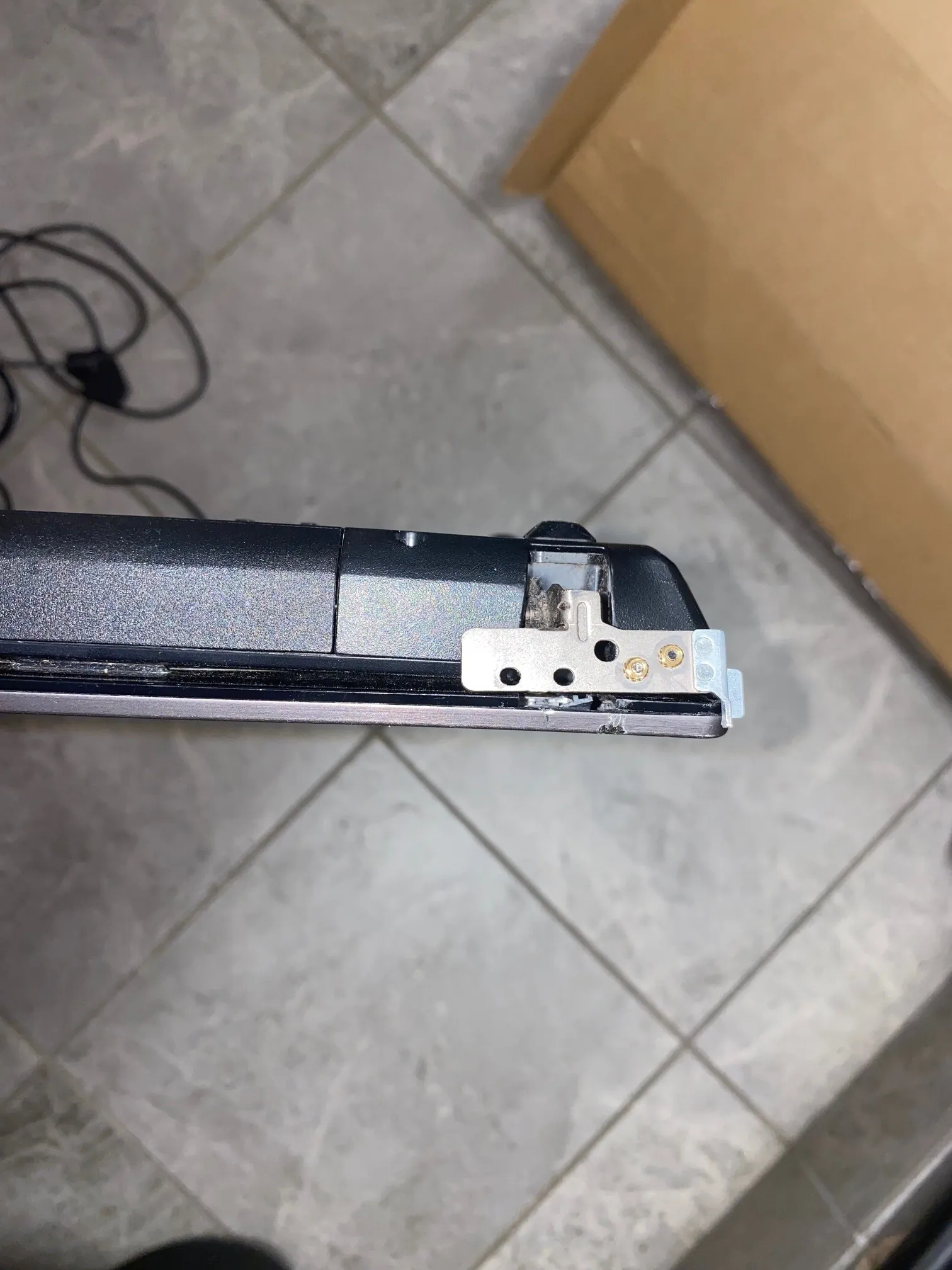 Zbliżenie na uszkodzoną krawędź czarnej obudowy urządzenia elektronicznego, prawdopodobnie laptopa. Widoczny jest ułamany fragment plastiku oraz wystający metalowy element mocujący z otworem na śrubę. Urządzenie leży na szarych, ceramicznych płytkach podłogowych. W tle widoczny jest fragment brązowego kartonu oraz czarny kabel z wtyczką.