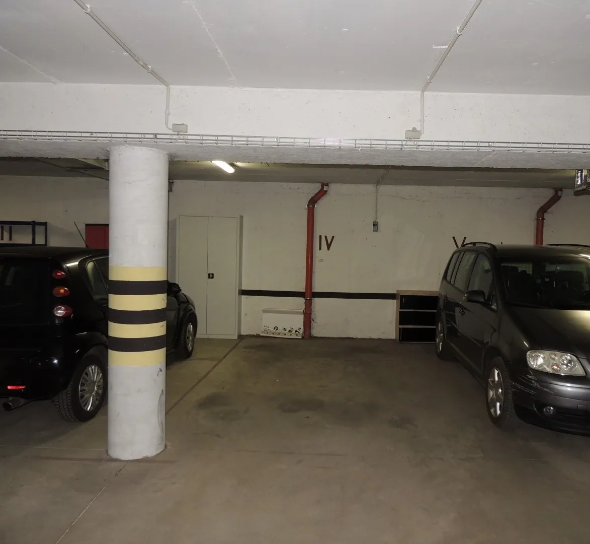 Podziemny parking z betonową podłogą i białymi ścianami. Po lewej stronie stoi czarny samochód osobowy, a obok niego znajduje się biały, okrągły filar z trzema poziomymi, żółtymi pasami. W tle widoczne są białe drzwi oraz rura kanalizacyjna w kolorze czerwonym, obok której znajduje się oznaczenie 'IV'. Po prawej stronie zaparkowany jest czarny samochód typu minivan.