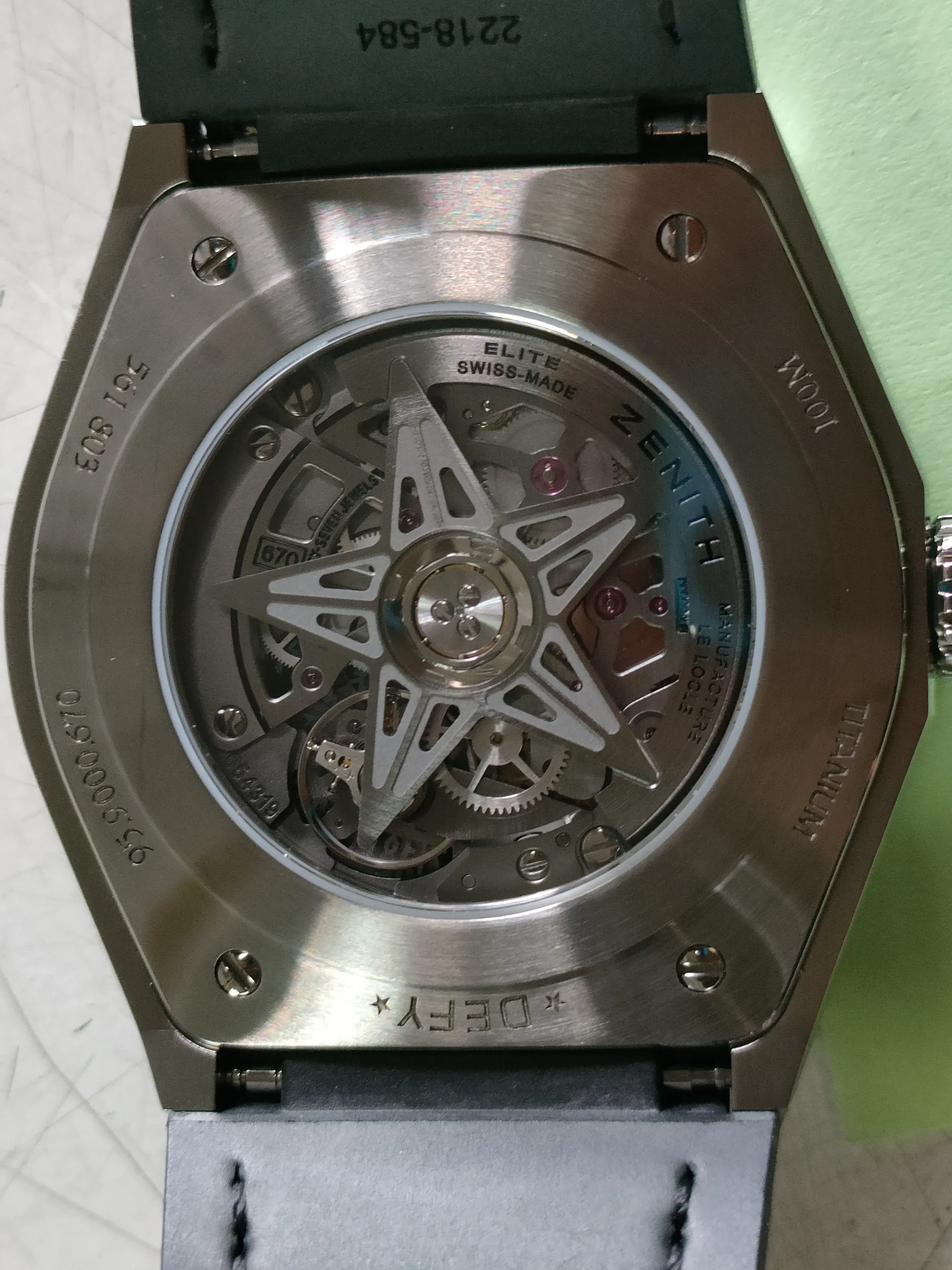 Zbliżenie na tylną część zegarka z widocznym mechanizmem. Metalowa obudowa z napisami: *DEFY*, ZENITH, ELITE SWISS-MADE, TITANIUM, 100M, 561 803, 95.9000.670, 2218-584 oraz inne numery seryjne. W centrum widoczny gwiaździsty rotor i zębatki mechanizmu. Pasek zegarka czarny z białymi szwami po lewej stronie, zielona powierzchnia w tle.