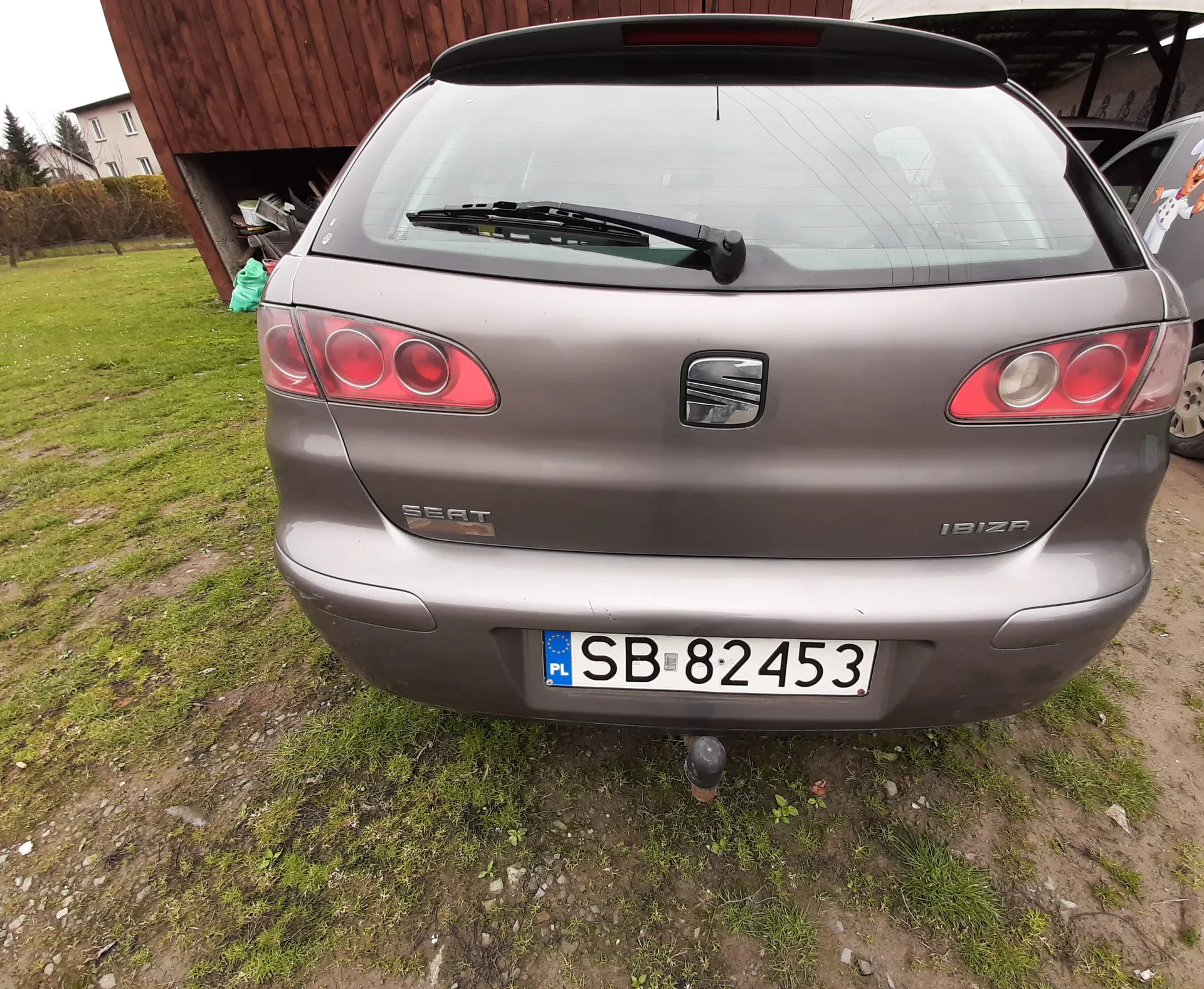 Tył szarego samochodu marki SEAT model Ibiza z tablicą rejestracyjną SB 82453 (PL). Samochód zaparkowany na trawiastym i częściowo błotnistym terenie. W tle drewniana budowla z przechowywanymi przedmiotami oraz fragment innego pojazdu z kreskówkowym motywem. Widoczne są czerwone światła tylne, logo marki oraz napisy "SEAT" i "IBIZA" na bagażniku.