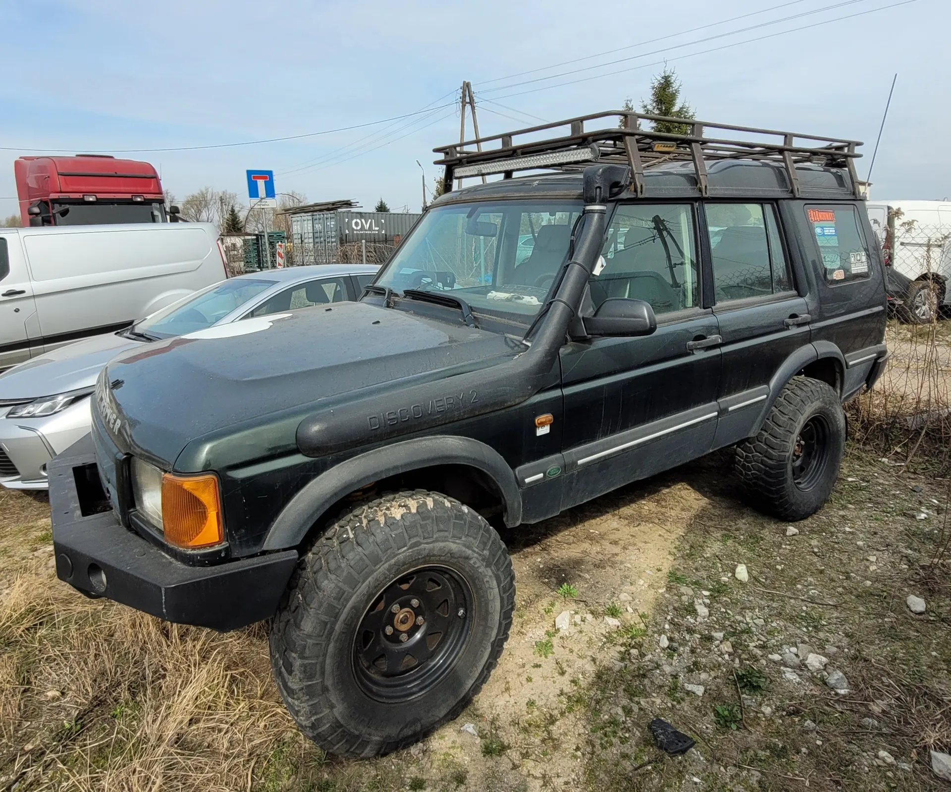 Ciemnozielony samochód terenowy Land Rover Discovery 2 z dachowym bagażnikiem i dużymi oponami terenowymi, zaparkowany na żwirowym obszarze z suchą trawą. W tle srebrny samochód osobowy, biały furgon i czerwony ciężarówka. Widoczny niebieski znak drogowy z białym 'T' oraz kontenery z napisem 'OVL'. Niebo częściowo zachmurzone, otoczenie przemysłowe z suchą trawą i kamieniami na ziemi.