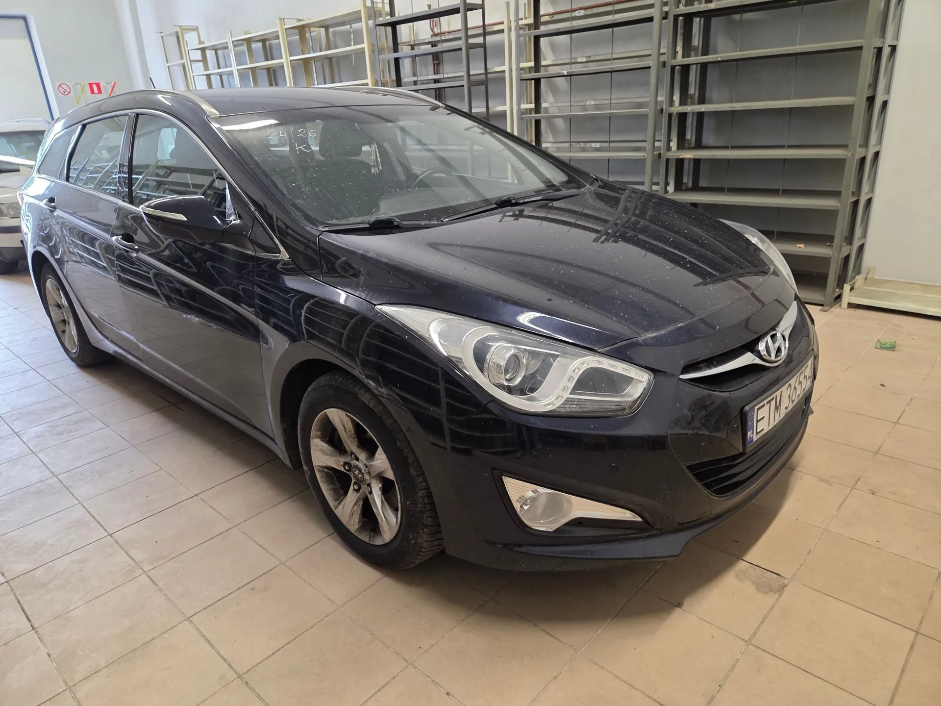 Czarny samochód osobowy marki Hyundai w nadwoziu kombi, zaparkowany wewnątrz pomieszczenia przypominającego magazyn lub warsztat z metalowymi regałami w tle. Pojazd jest zakurzony, widoczna jest tablica rejestracyjna o numerze ETN 3655Z.
