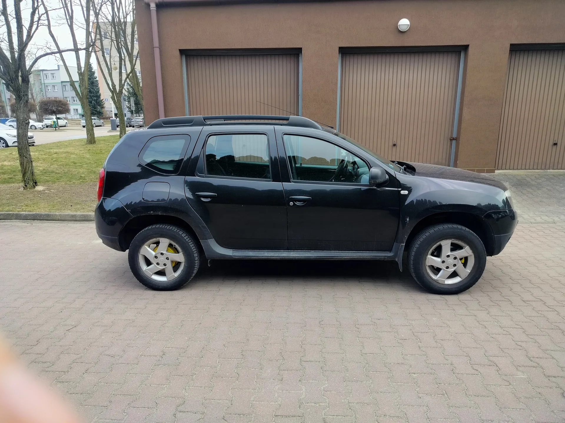 Czarny SUV zaparkowany na wybrukowanym placu przed brązowym budynkiem z trzema zamkniętymi bramami garażowymi. Pojazd posiada srebrne felgi i dachówkę. W tle widoczne są bezlistne drzewa, trawnik oraz inne budynki. Niebo jest pochmurne.