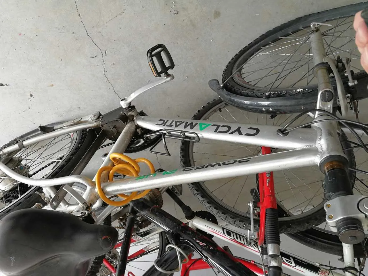 Srebrny rower z napisem 'CYCLAMATIC' i 'POWER' na ramie, z żółtym zwojonym kablem zabezpieczającym. W tle widoczne są inne rowery, w tym czerwony i biały. Podłoga z betonu, widoczna ręka w lewym górnym rogu.