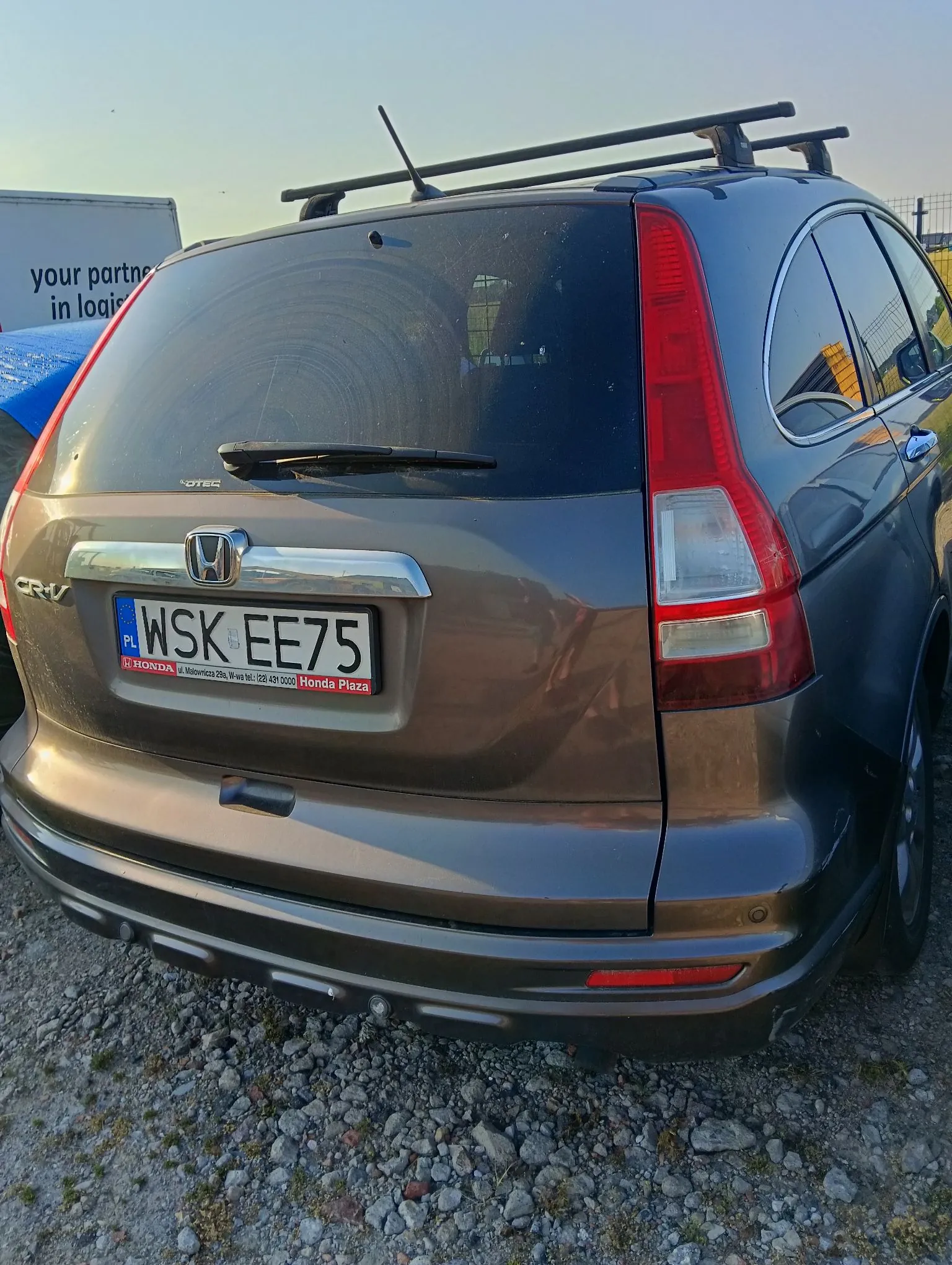 Tylny widok brązowego samochodu marki Honda CR-V zaparkowanego na żwirowym podłożu. Na klapie bagażnika znajduje się polska tablica rejestracyjna o numerze WSK EE75 oraz logo Hondy. Poniżej tablicy widnieje napis: 'HONDA ul. Małowianki 29a, Wola Śląska tel. / 020 / 429 0000 Honda Plaza'. Na dachu samochodu zamontowany jest czarny bagażnik dachowy. W tle po lewej stronie widoczny jest fragment białej naczepy z napisem 'your partner in logis'.