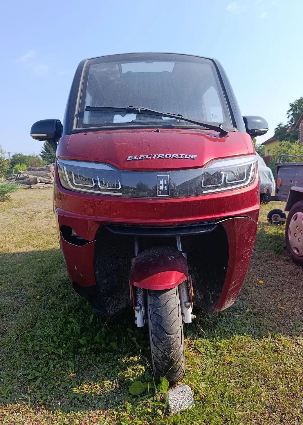 Widok z przodu czerwonego skutera elektrycznego marki ELECTRORIDE, zaparkowanego na trawie. Pojazd ma nowoczesny design z szerokimi reflektorami LED i czarną szybą przednią. Na masce widnieje biały napis ELECTRORIDE. Przednie koło jest skierowane prosto, a pod nim znajduje się mały biały kamień.