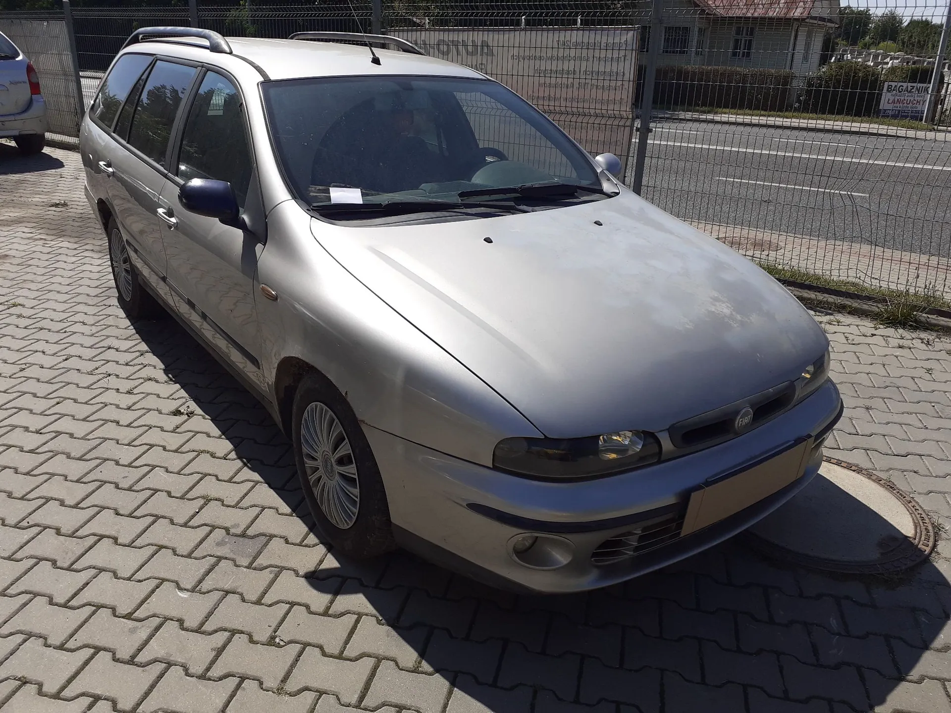 Srebrny samochód osobowy typu kombi marki Fiat zaparkowany na kostce brukowej. Pojazd jest ustawiony pod kątem, widoczny z przodu i z lewej strony. W tle znajduje się metalowy płot z siatki oraz fragment drogi.