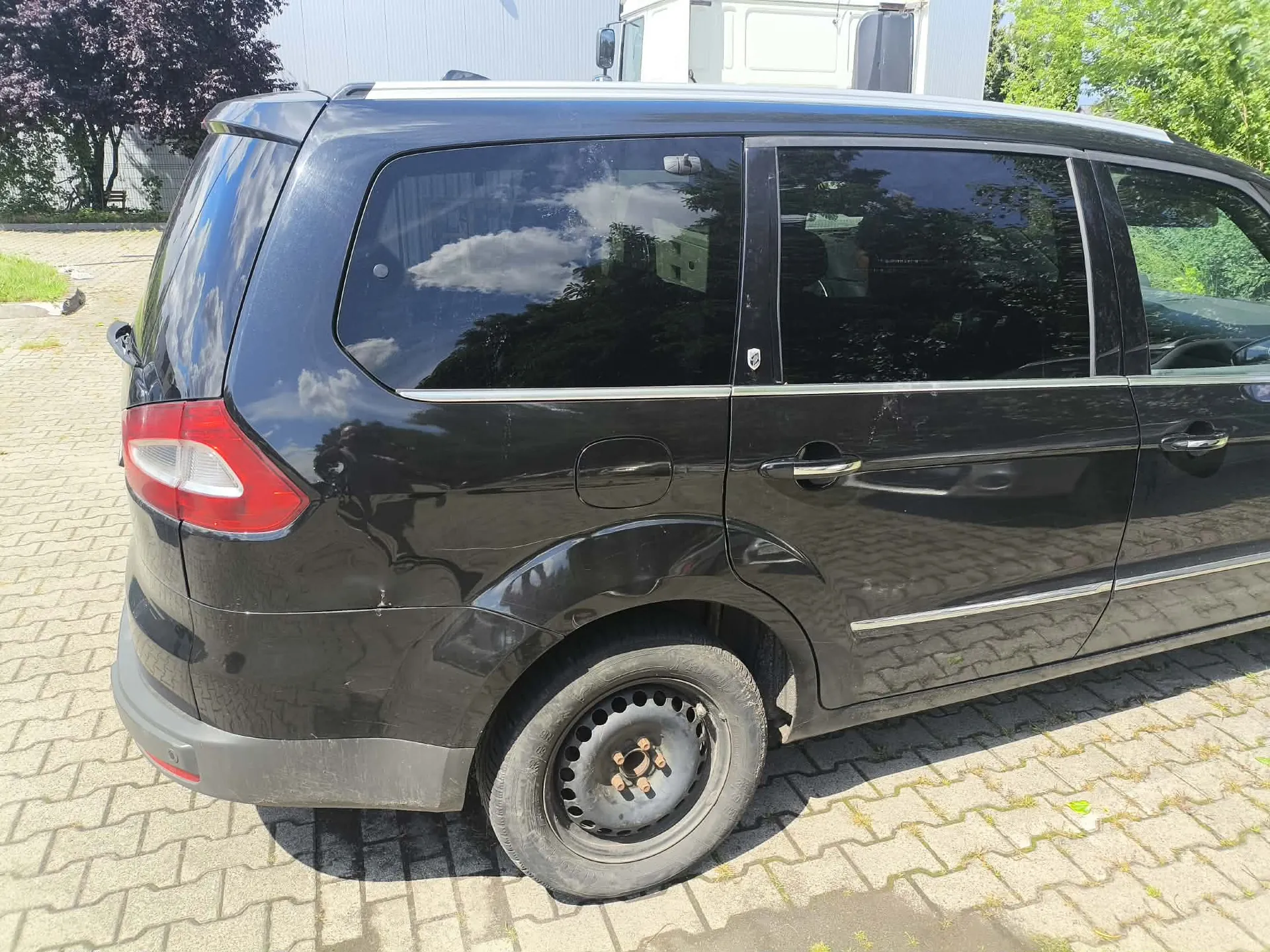 Czarny samochód typu minivan zaparkowany na kostce brukowej. Zdjęcie przedstawia tylną i boczną część pojazdu. Samochód ma przyciemniane szyby i stalowe felgi bez kołpaków.