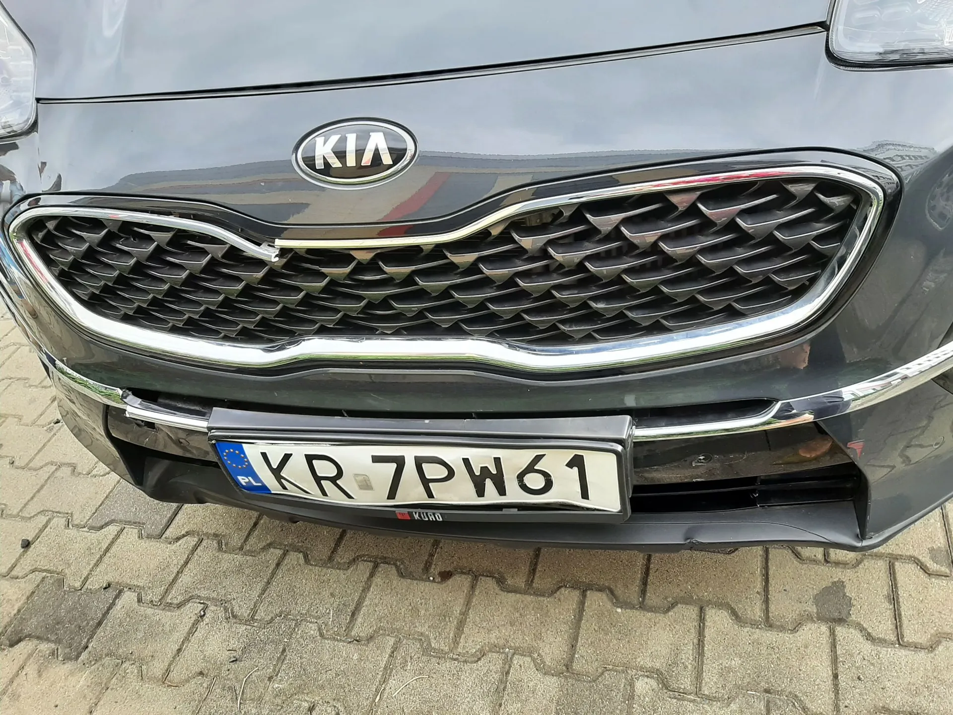 Zbliżenie na przód ciemnoszarego samochodu marki Kia. Widoczny jest chromowany grill o strukturze plastra miodu, logo KIA nad grillem oraz polska tablica rejestracyjna o numerze KR 7PW61. Samochód stoi na kostce brukowej.