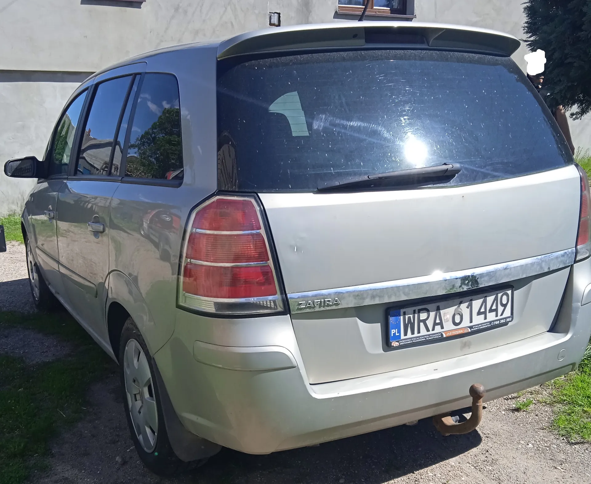 Tył srebrnego samochodu Opel Zafira zaparkowanego na żwirowym podjeździe. Widoczna tablica rejestracyjna PL WRA 61449 z dodatkowym napisem na dole, czerwone światła tylnego, napis ZAFIRA na bagażniku oraz hak holowniczy. W tle budynek z oknem i drzewo. Samochód ma lekko zabrudzoną karoserię, a na szybie tylnej odbijają się drzewa i niebo.