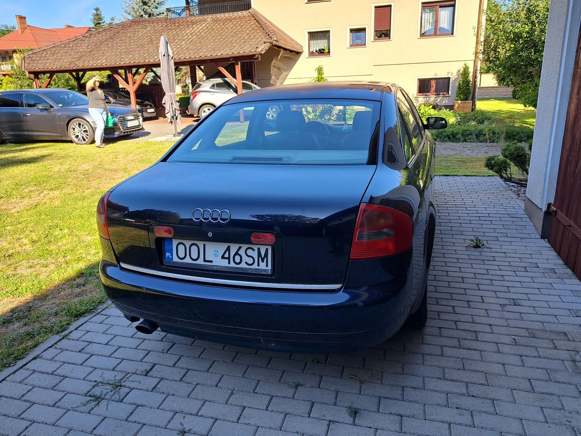 Tylny widok ciemnoniebieskiego samochodu marki Audi zaparkowanego na kostce brukowej. Samochód ma polską tablicę rejestracyjną o numerze OOL 46SM. W tle widoczny jest dom z żółtą elewacją, zadaszenie nad podjazdem, pod którym stoi srebrny samochód, oraz szary samochód zaparkowany na trawniku, przy którym stoi kobieta. Wokół znajduje się zielona trawa i krzewy.
