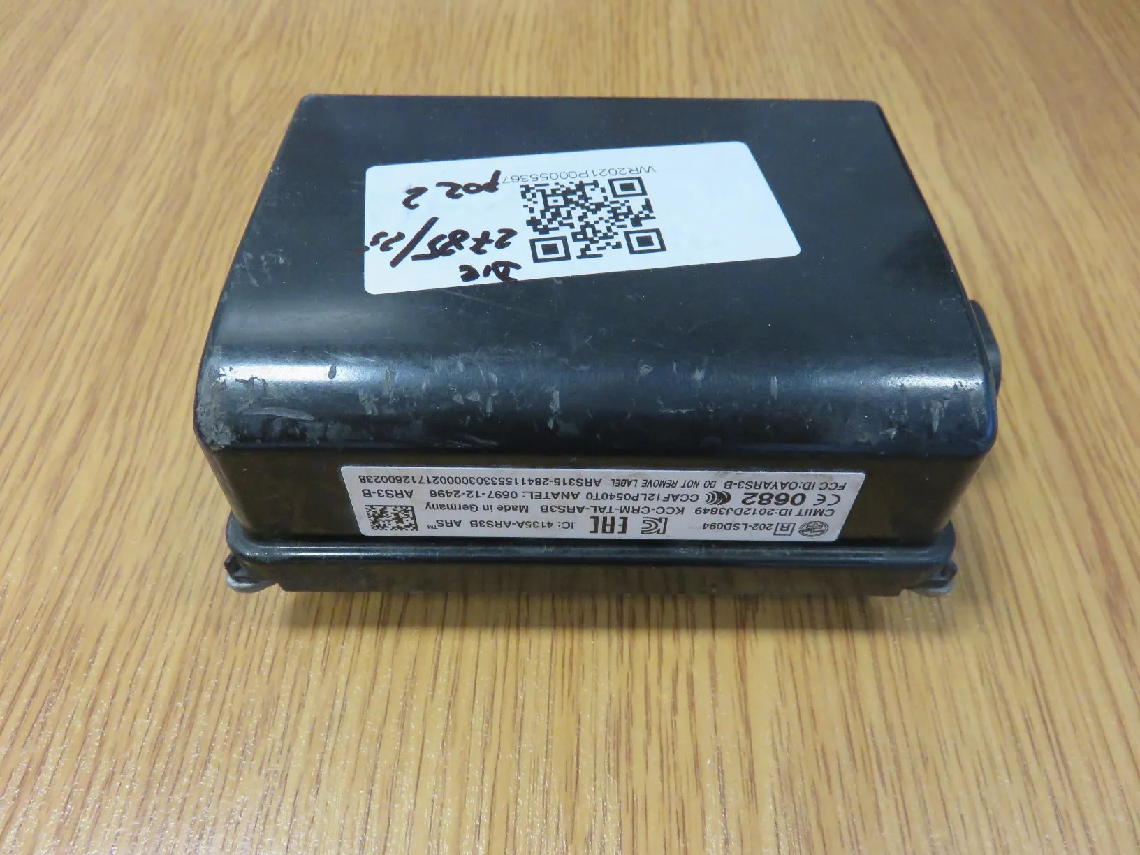 Czarny, prostokątny moduł elektroniczny leżący na drewnianej powierzchni. Na górnej obudowie znajduje się biała naklejka z kodem QR, kodami kreskowymi oraz odręcznymi zapisami. Na bocznej krawędzi widnieją białe napisy techniczne, w tym: 'CE 0682', 'FCC ID: OAYARS3-B', 'IC: 4135A-ARS3B', 'Made in Germany' oraz inne oznaczenia certyfikatów i numery seryjne.