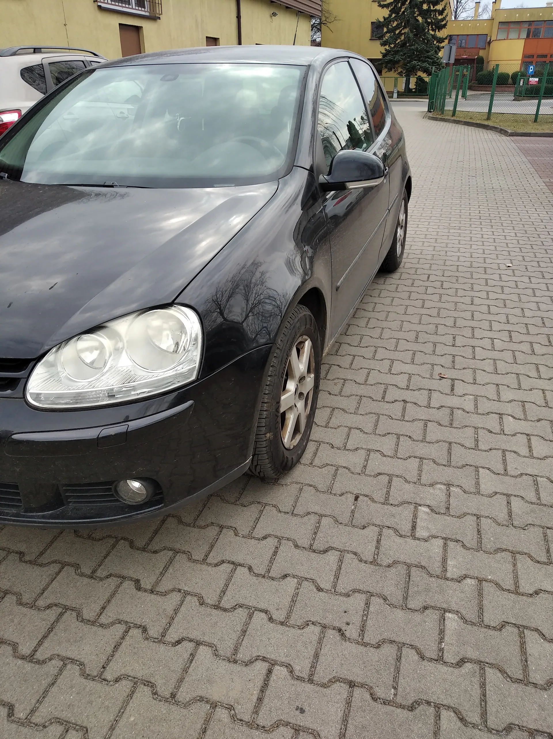 Czarny samochód osobowy zaparkowany na brukowanej powierzchni. W tle żółty budynek, zielone ogrodzenie, drzewo oraz częściowo widoczny biały samochód. Na drodze widoczny znak parkingowy z literą P.