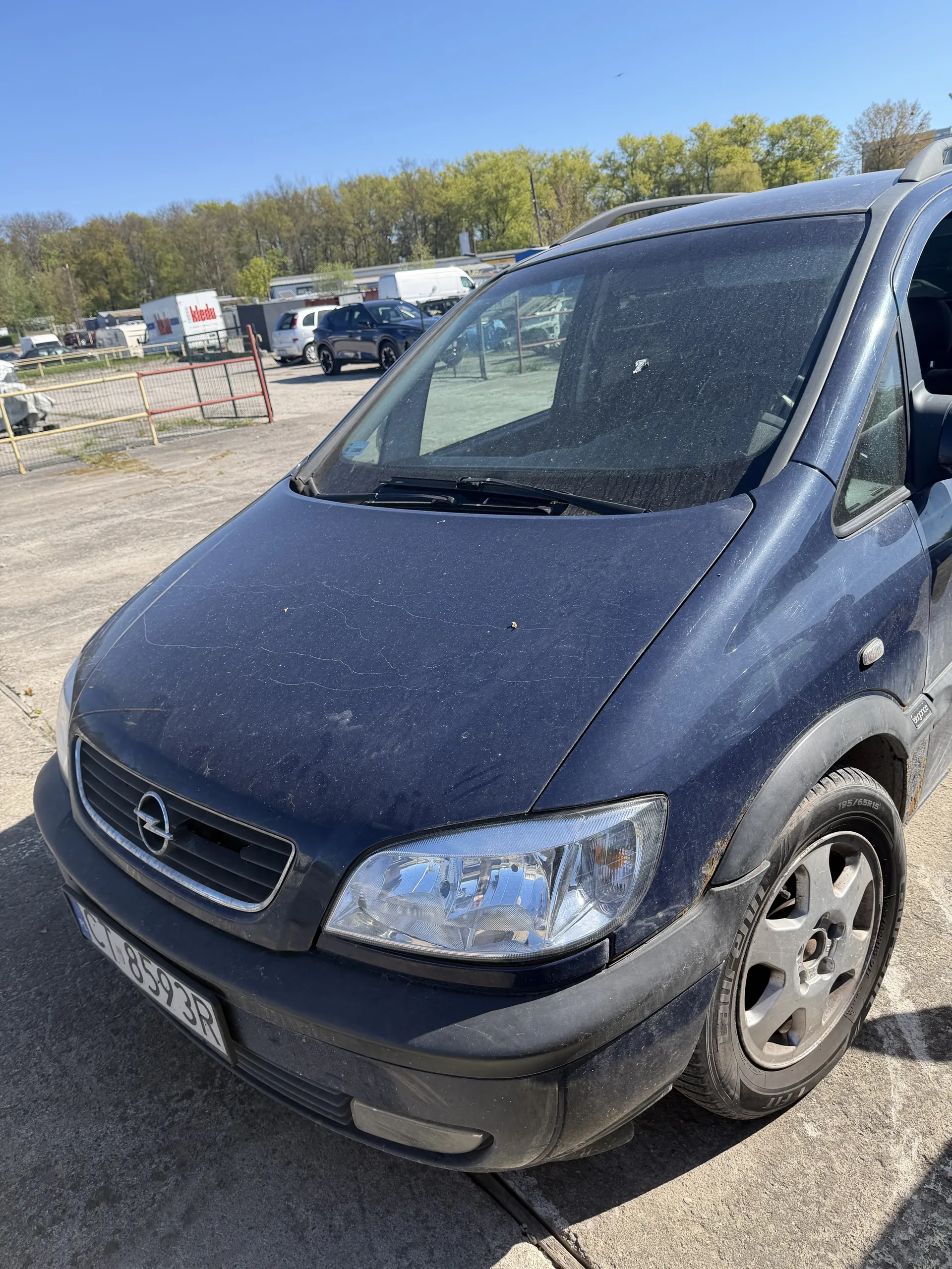Ciemnoniebieski samochód marki Opel z widocznym logo na atrapie chłodnicy. Na przednim zderzaku zamontowano zapasowe koło. Tablica rejestracyjna z numerem CT 8593R. Na masce widoczne są drobne zadrapania. W tle parking z innymi pojazdami, ogrodzeniem, drzewami i jasnym niebem.