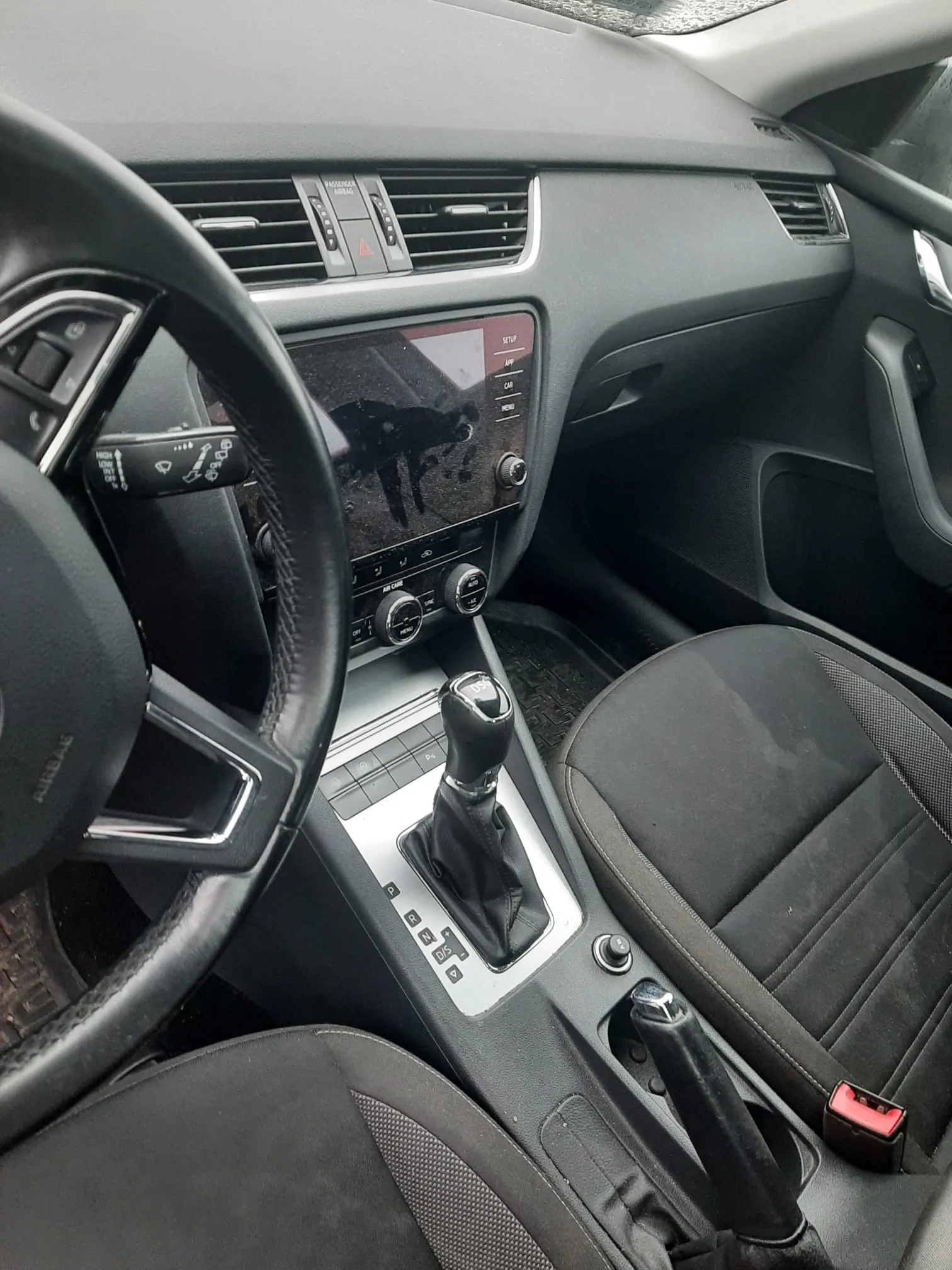 Wnętrze samochodu z widocznym kierownicą po lewej stronie, czarnym deski rozdzielczej z wentylacjami, ekranem centralnym z odbiciem, dźwignią zmiany biegów, czarnymi siedzeniami z siateczkowym wzorem oraz czerwonym zapięciem pasa bezpieczeństwa na siedzeniu pasażera. Na desce rozdzielczej widoczne są przyciski i pokrętła sterujące systemem audio oraz klimatyzacją.
