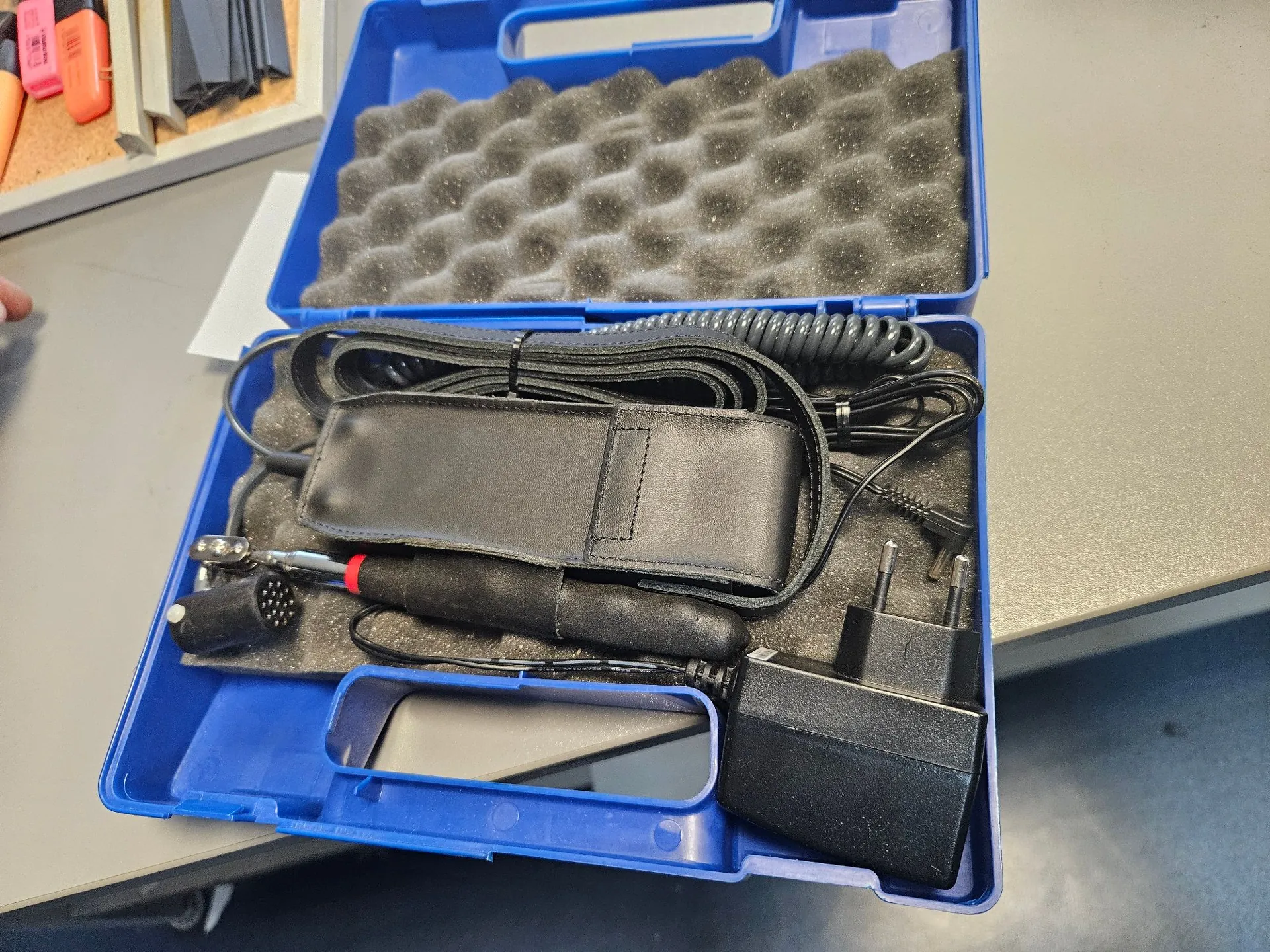 Otwarta niebieska plastikowa walizka transportowa z wkładem z ciemnoszarej pianki. Wewnątrz znajdują się: czarny zasilacz z wtyczką sieciową, czarne etui z ekoskóry, zwinięty czarny kabel oraz urządzenie z czarną rączką i metalowym złączem wielopinowym.