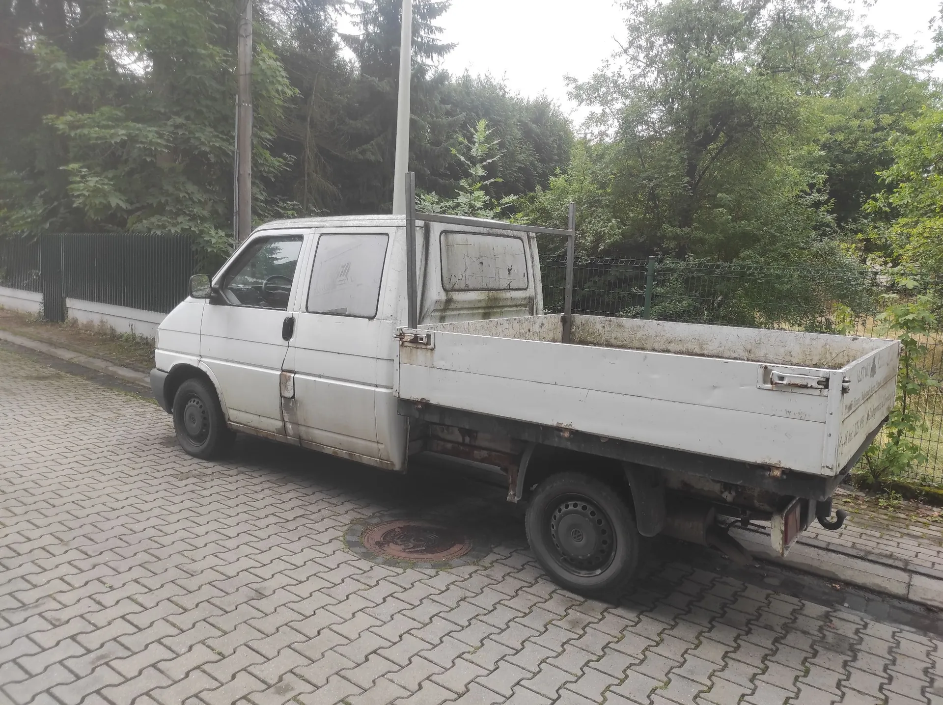 Biały samochód dostawczy z otwartą paką, zaparkowany na kostce brukowej. Pojazd stoi na tle gęstych, zielonych drzew i metalowego ogrodzenia.