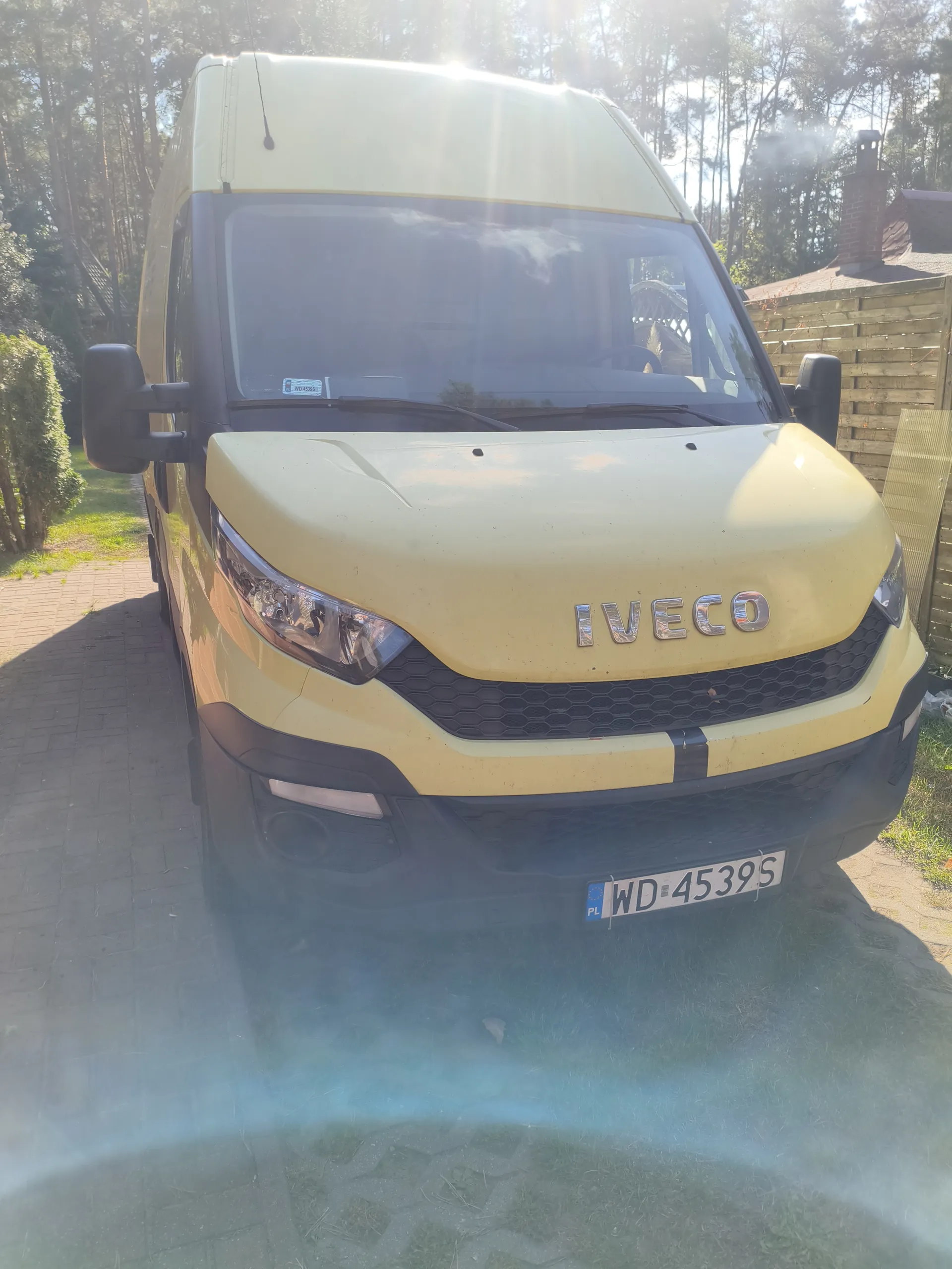 Żółty samochód dostawczy Iveco z tablicą rejestracyjną WD-4539S (PL) zaparkowany na brukowanej powierzchni. W tle drzewa, drewniany płot i dom z ceglanym kominkiem. Słoneczna pogoda, widoczne promienie słońca.