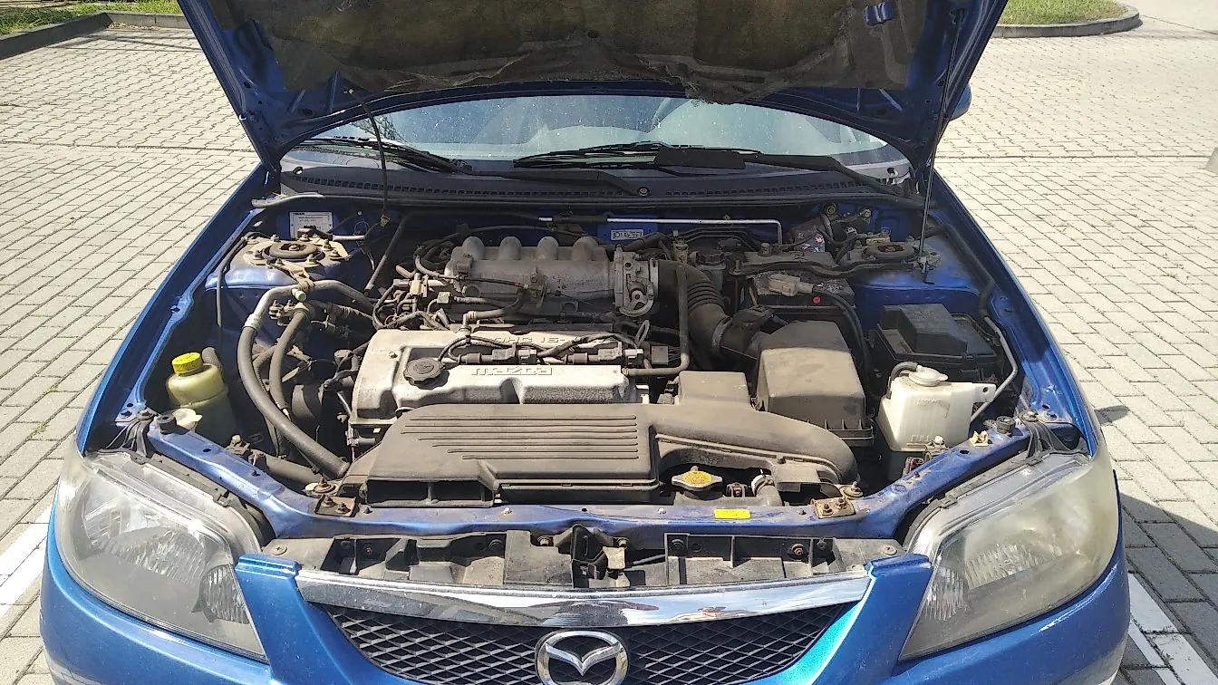 Widok z góry na otwartą maskę niebieskiego samochodu marki Mazda, ukazujący komorę silnika z widocznymi elementami mechanicznymi, przewodami, akumulatorem oraz zbiornikiem płynu wycieraczek. W dolnej części zdjęcia widoczny jest fragment przedniego grilla z logo Mazdy oraz reflektory.