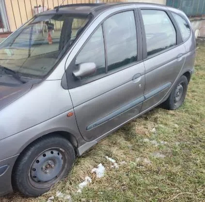 Szary samochód osobowy typu minivan zaparkowany na trawie z resztkami śniegu. Pojazd ma czarne felgi, a na boku widoczny jest niewielki napis. W tle znajduje się budynek z oknem i drzwiami. Trawa jest częściowo zasłonięta śniegiem, a samochód stoi pod kątem do obserwatora.