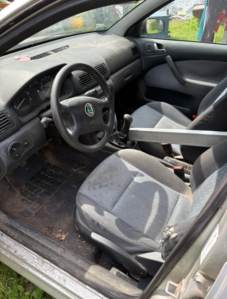 Wnętrze samochodu od strony kierowcy. Widoczna jest zakurzona deska rozdzielcza, kierownica z logo marki Škoda, fotele z szarej tkaniny oraz gumowe dywaniki. Drzwi pasażera są otwarte, a wewnątrz auta znajduje się metalowy element konstrukcyjny przebiegający poziomo nad fotelem.