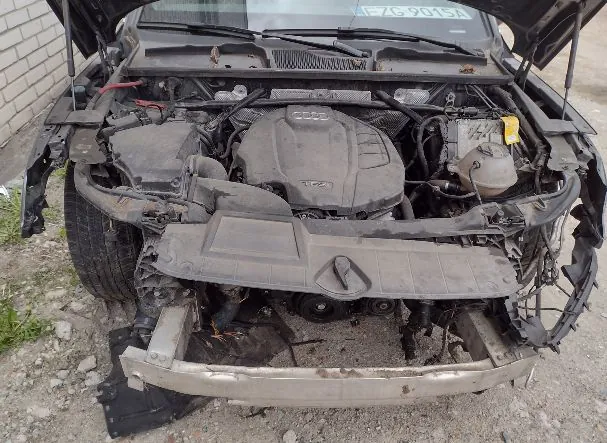 Zdjęcie przodu uszkodzonego, czarnego samochodu marki Audi z otwartą maską. Brak przedniego zderzaka i reflektorów, odsłaniając silnik oraz metalowy wzmocniony element konstrukcyjny zderzaka. Na przedniej szybie widoczna jest tablica rejestracyjna o numerze EZG 9015A.
