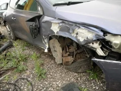 Zniszczony szary samochód osobowy z uszkodzonym przodem. Widoczne są zdeformowane elementy karoserii, brak przedniego koła oraz odsłonięte podzespoły mechaniczne. Samochód stoi na podłożu z żwiru i trawy, w tle częściowo widoczny inny pojazd.