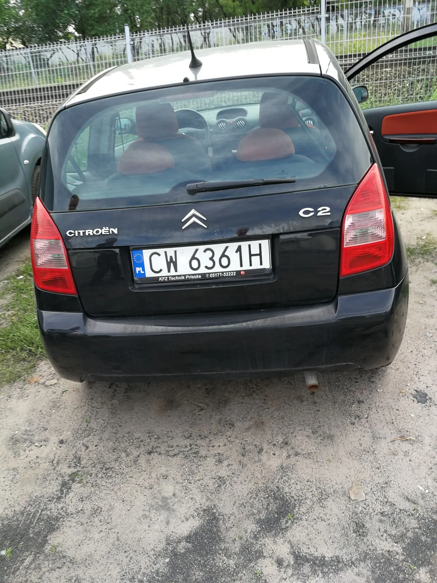 Tylny widok czarnego samochodu marki Citroën model C2. Pojazd ma polską tablicę rejestracyjną o numerze CW 6361H. Na klapie bagażnika widoczne są napisy 'CITROËN' oraz 'C2' oraz logo marki. Wewnątrz samochodu widoczne są brązowe zagłówki foteli. Samochód stoi na piaszczystym podłożu przed metalowym ogrodzeniem.