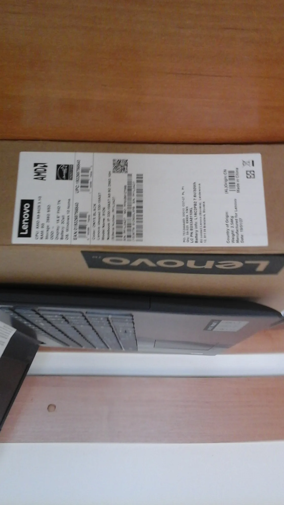 Pudełko laptopa Lenovo z widoczną etykietą zawierającą specyfikację techniczną, logotypami Lenovo i AMD, kodami kreskowymi oraz kodem QR. Część laptopa z klawiaturą i touchpadem widoczna po lewej stronie. Tło z drewnianą powierzchnią.