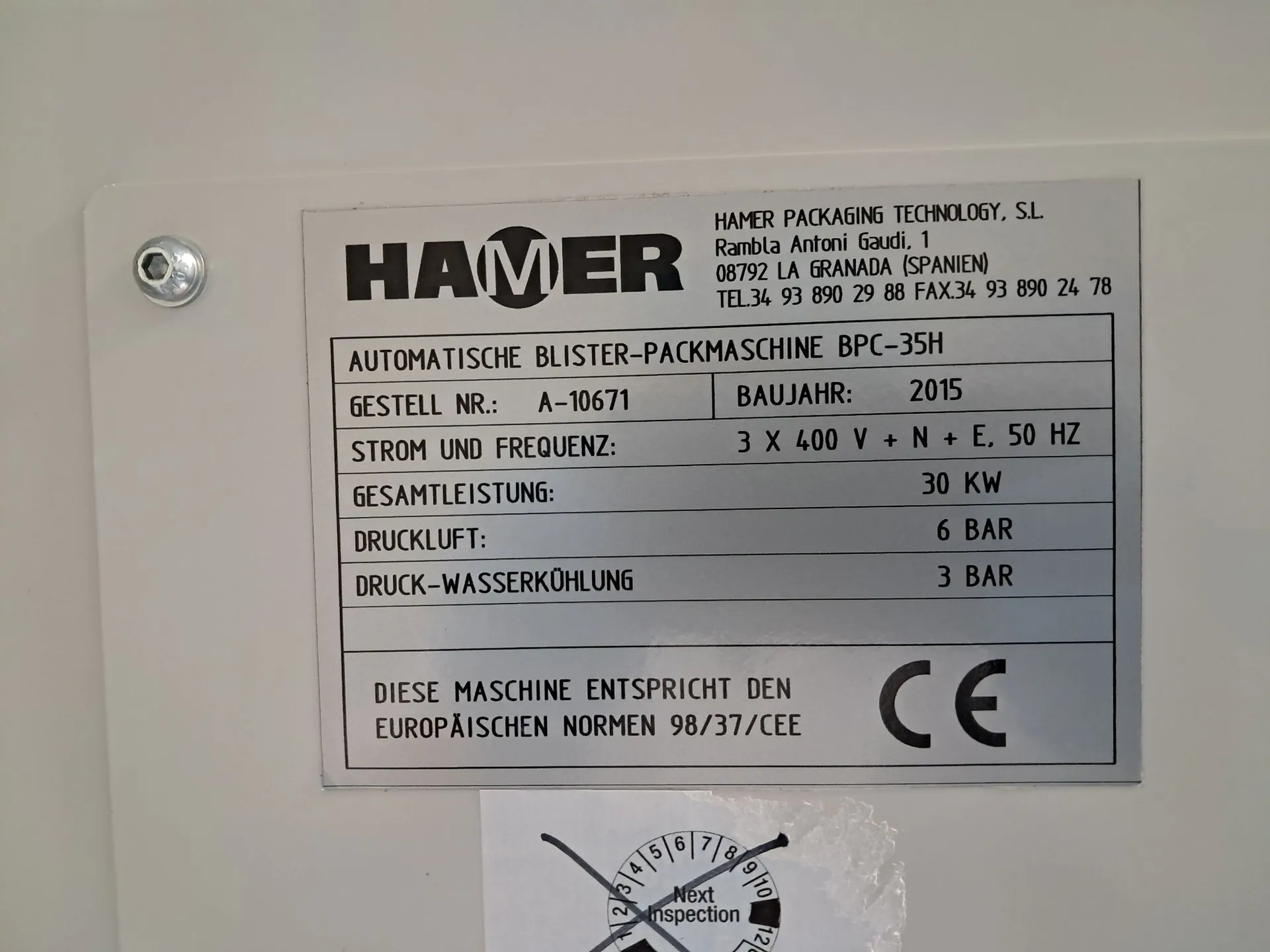 Srebrna tabliczka znamionowa przymocowana do białej obudowy maszyny. Na górze znajduje się logo 'HAMER' oraz dane firmy: 'HAMER PACKAGING TECHNOLOGY, S.L. Rambla Antoni Gaudi, 1 08792 LA GRANADA (SPANIEN) TEL. 34 93 890 29 88 FAX. 34 93 890 24 78'. Poniżej znajduje się tabela z danymi technicznymi: 'AUTOMATISCHE BLISTER-PACKMASCHINE BPC-35H', 'GESTELL NR.: A-10671', 'BAUJAHR: 2015', 'STROM UND FREQUENZ: 3 X 400 V + N + E, 50 HZ', 'GESAMTLEISTUNG: 30 KW', 'DRUCKLUFT: 6 BAR', 'DRUCK-WASSERKÜHLUNG: 3 BAR'. Na dole tabliczki widnieje napis 'DIESE MASCHINE ENTSPRICHT DEN EUROPÄISCHEN NORMEN 98/37/CEE' oraz znak CE. Pod tabliczką znajduje się okrągła naklejka inspekcyjna z napisem 'Next Inspection'.