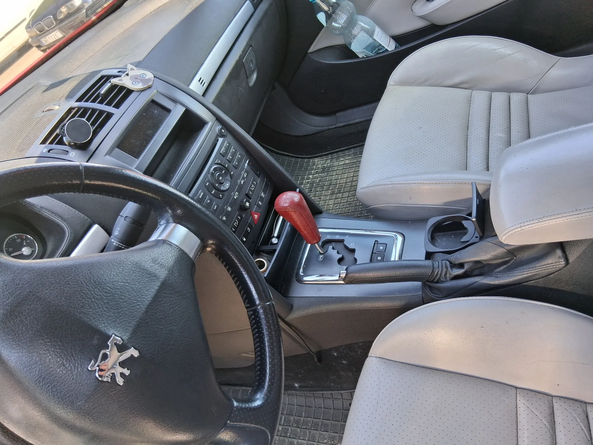 Wnętrze samochodu z widocznym kierownicą z logo Peugeot, czerwonym dźwignią zmiany biegów, jasnoszarymi perforowanymi fotelami skórzonymi, deska rozdzielcza z przyciskami i wentylacją, małym naklejką w kształcie kota na desce, butelką wody w kieszeni drzwi oraz częścią innego pojazdu widoczną przez przednią szybę.