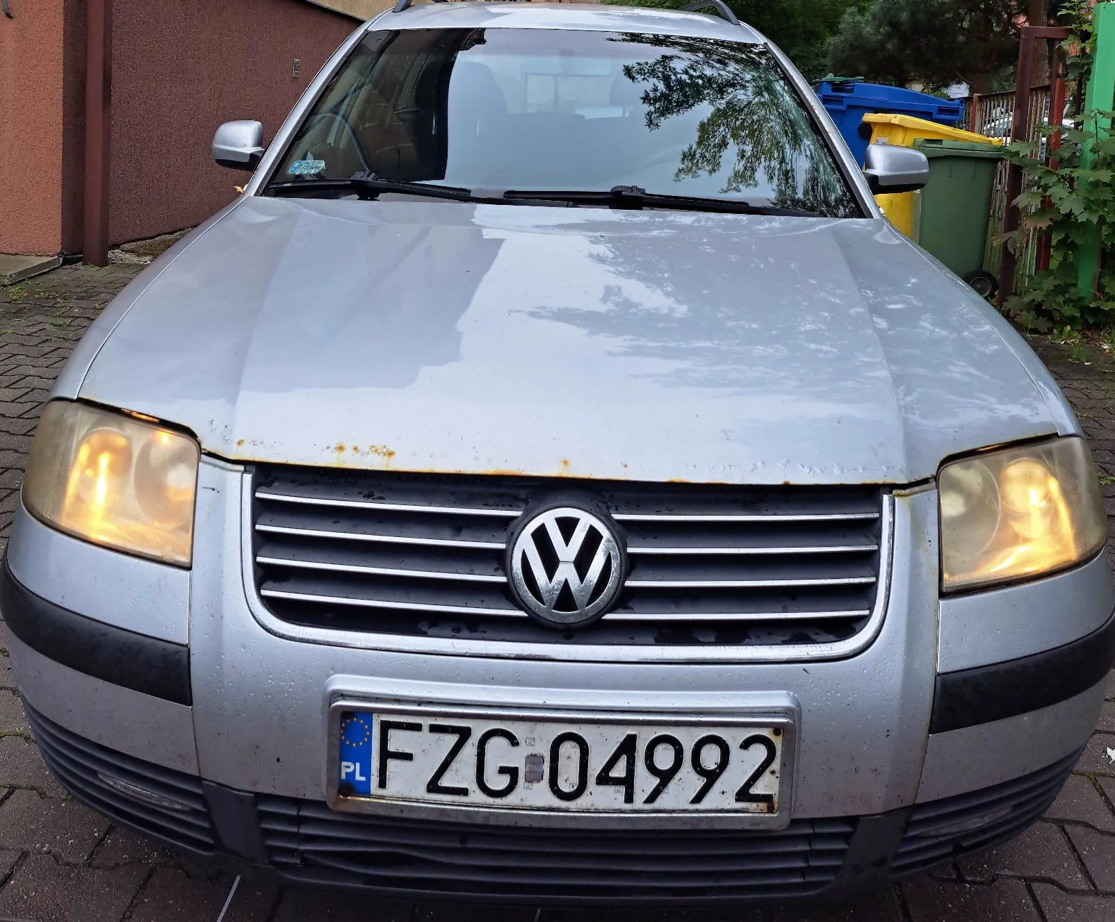 Srebrny samochód marki Volkswagen widziany od przodu. Pojazd ma włączone przednie światła, a na masce widoczne są ślady rdzy nad grillem. Samochód posiada polską tablicę rejestracyjną o numerze FZG 04992.