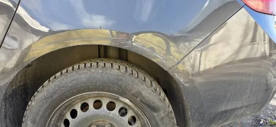 Zbliżenie na tylną część ciemnego samochodu, pokazujące koło z oponą o widocznych śladach zużycia i felgą z otworami. Nad kołem widoczna zabrudzona karoseria z odbiciami budynków i nieba. Tło stanowi drobny żwir.